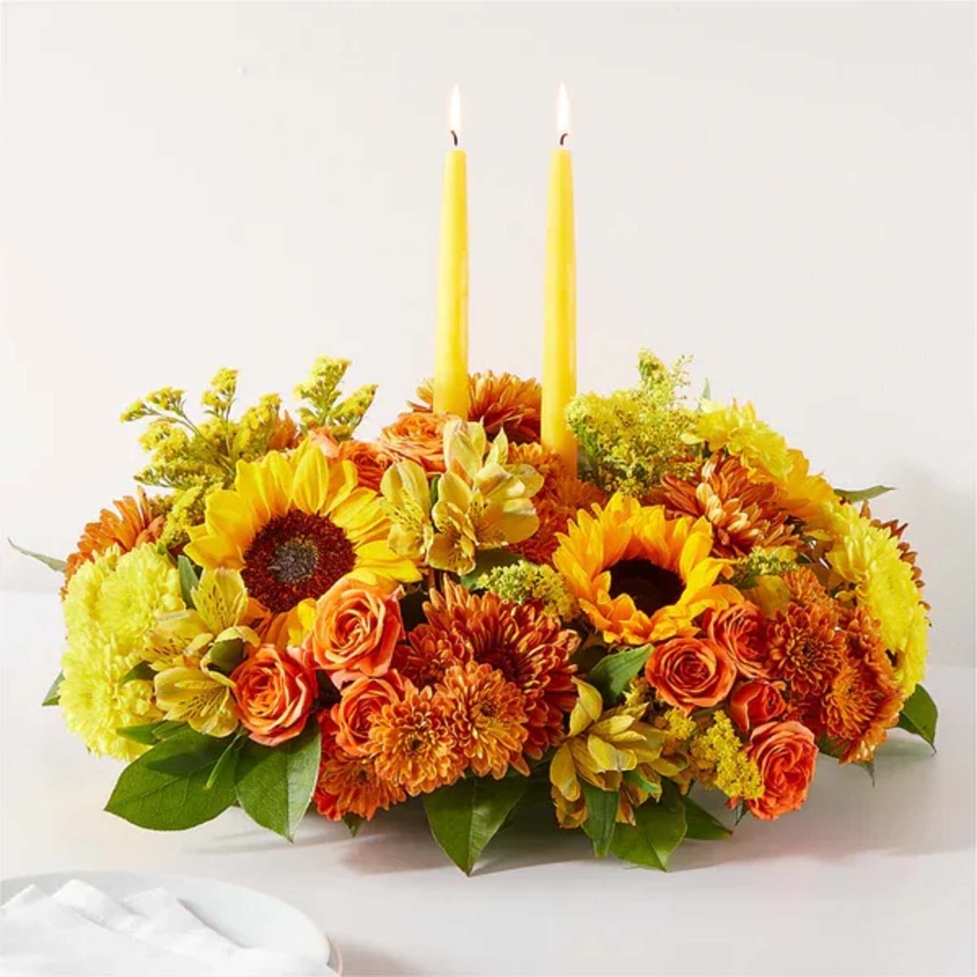 Yellow candles rise from a low arrangement of sunflowers, roses, and mums