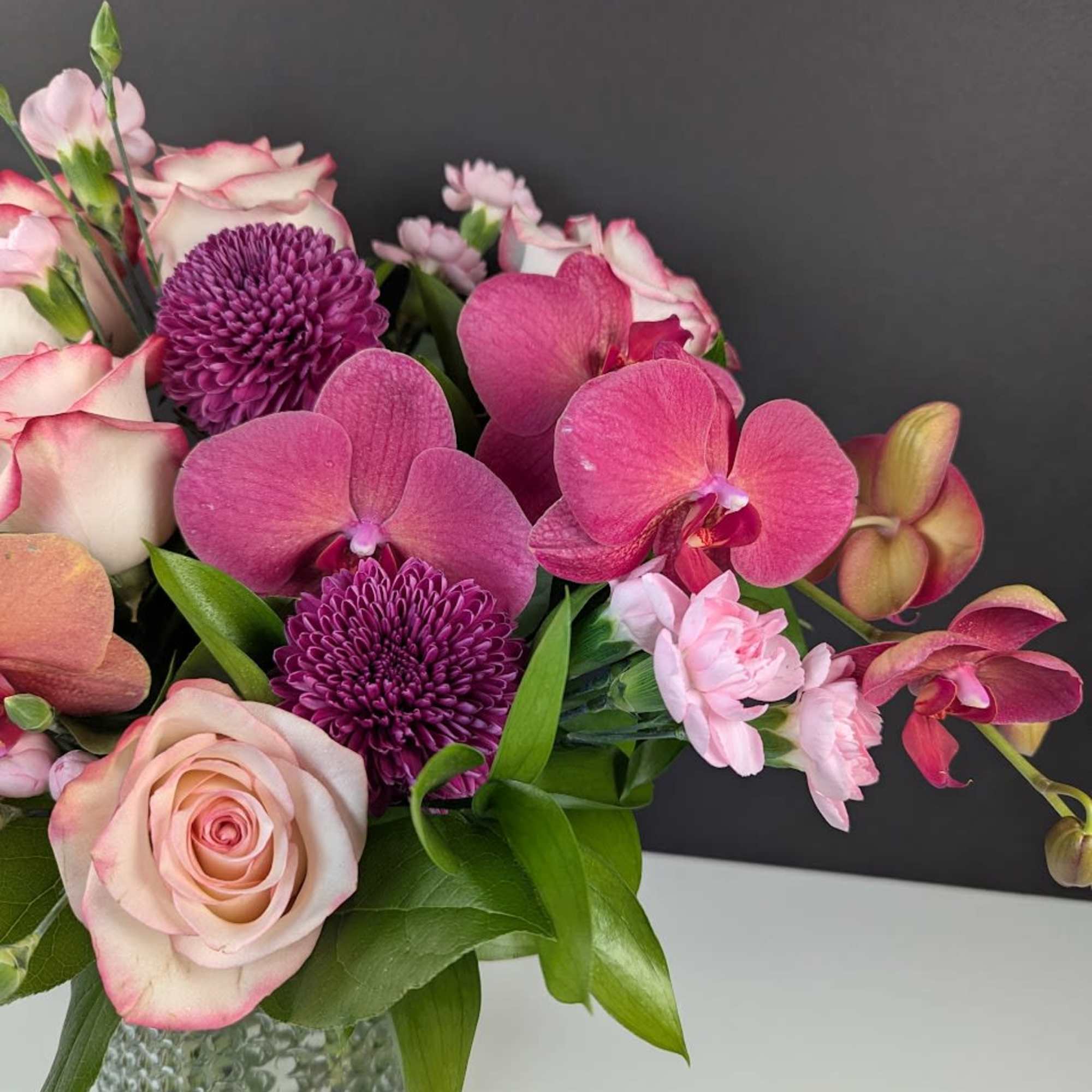 Compact arrangement of pink orchids, roses, and purple mums in a textured glass vase