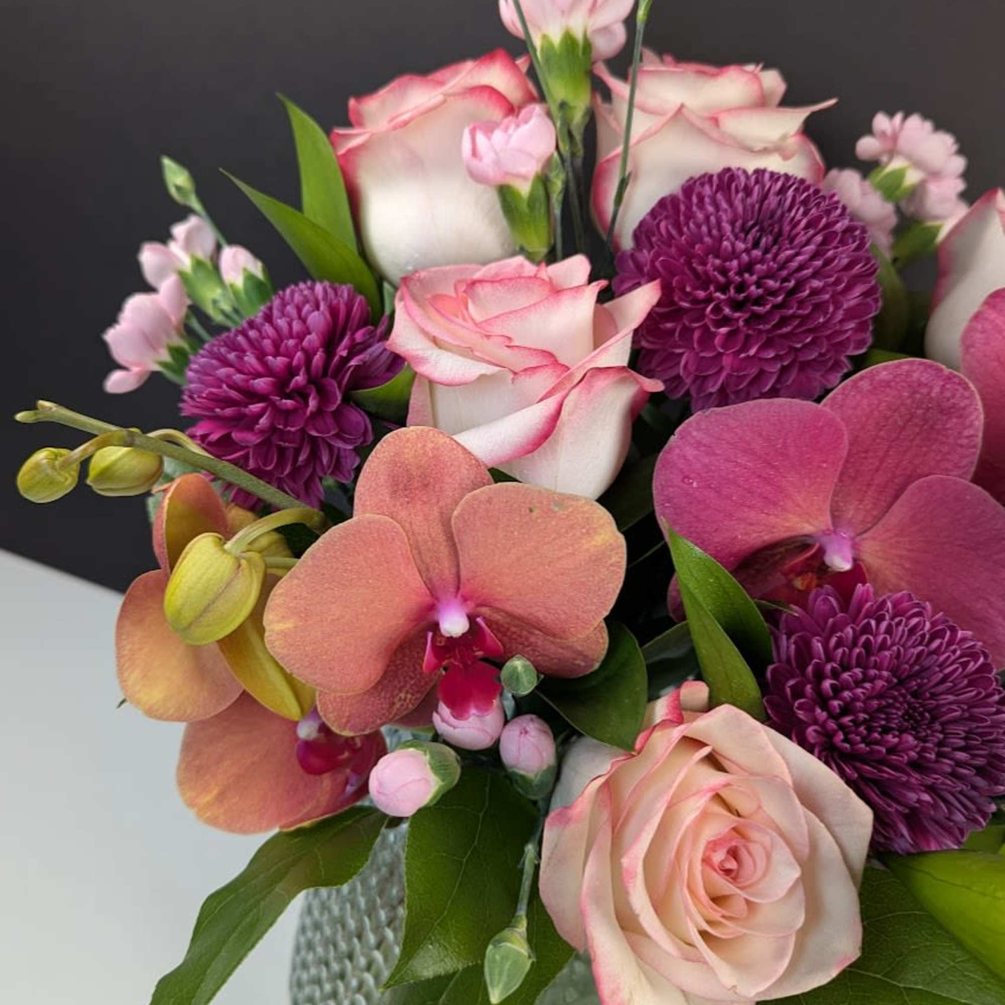 Compact bouquet of pink roses, purple mums, and peach orchids in a textured glass vase