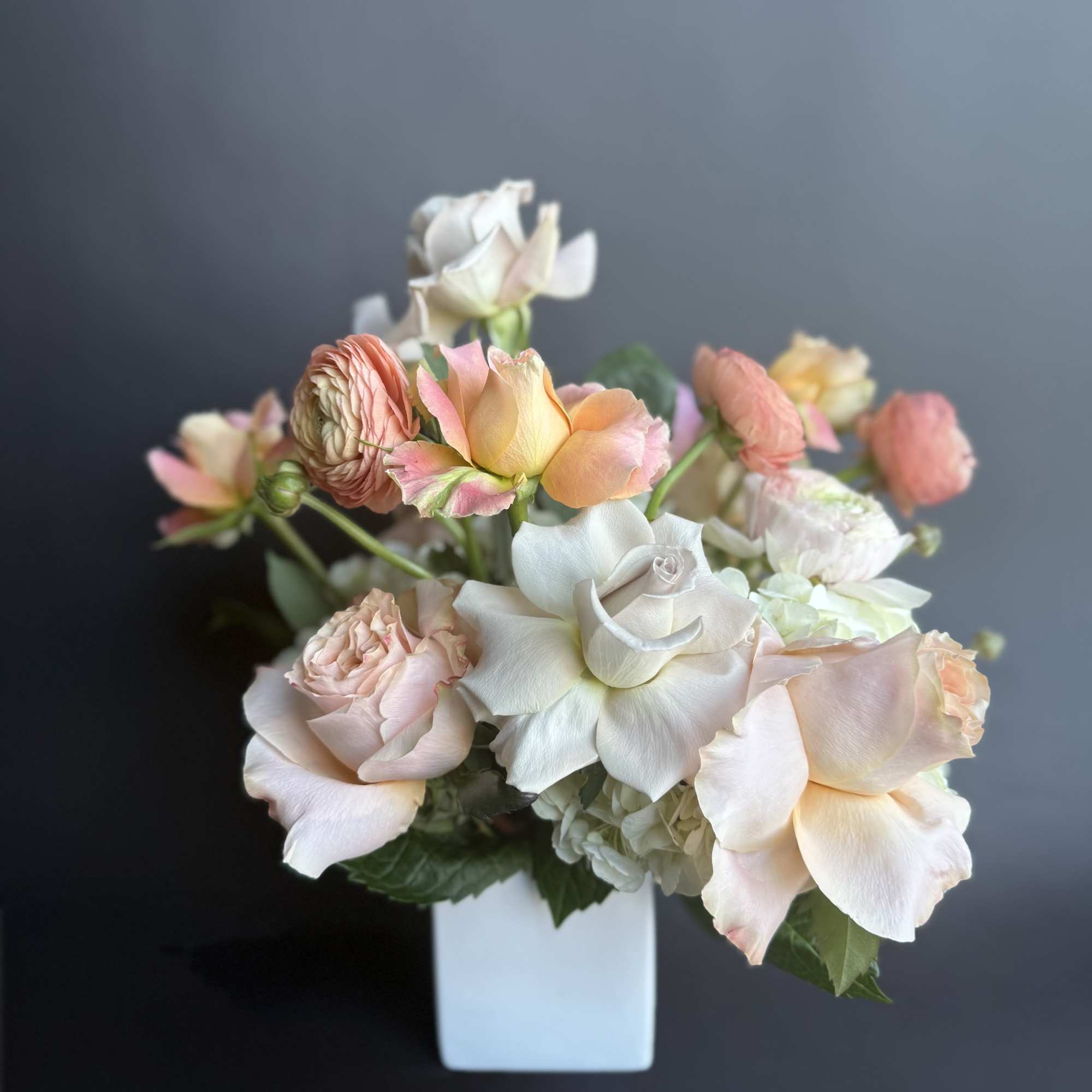 Low arrangement of blush and peach roses with ranunculus in a white cube vase