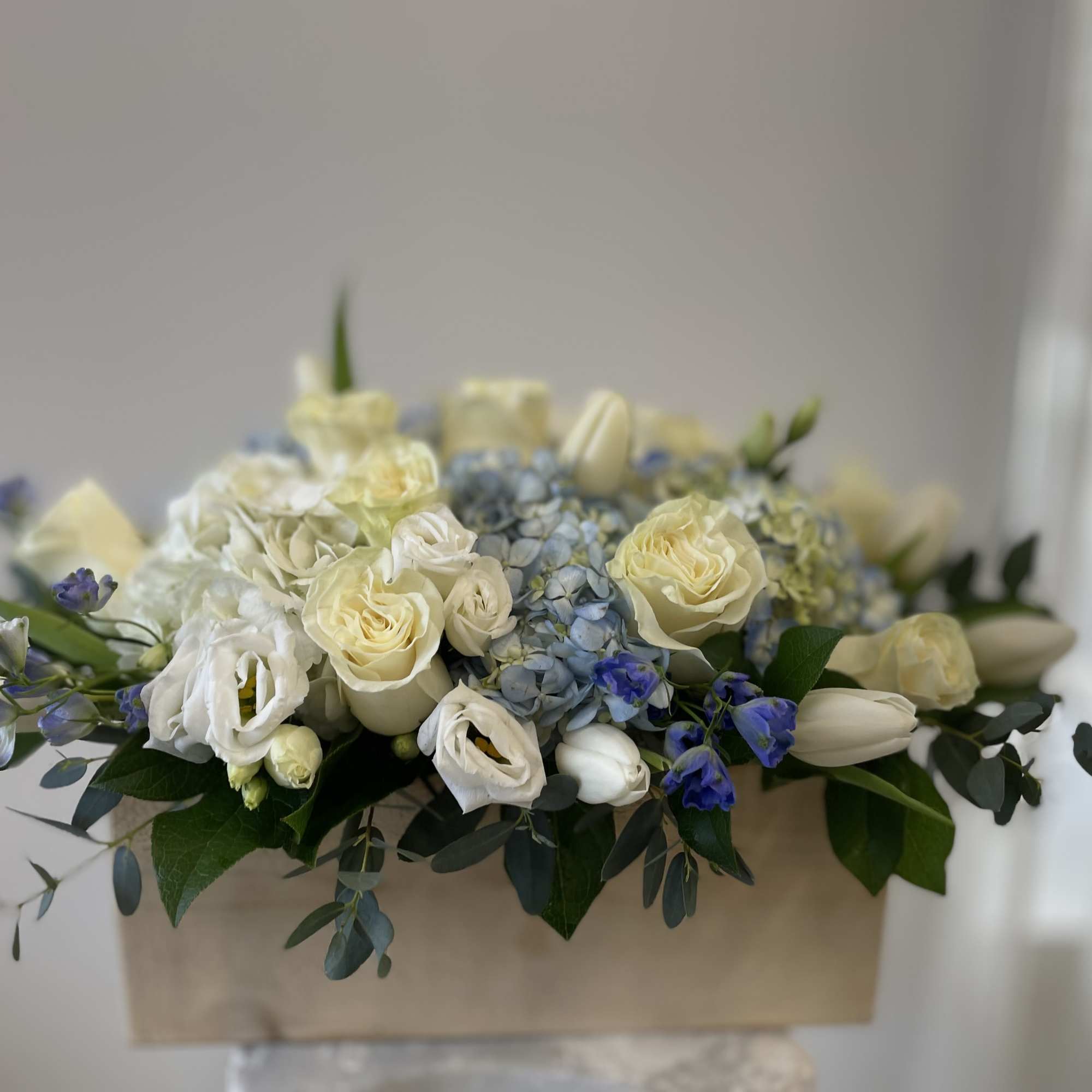 Low arrangement of cream roses, white tulips, and blue hydrangeas in a wooden box