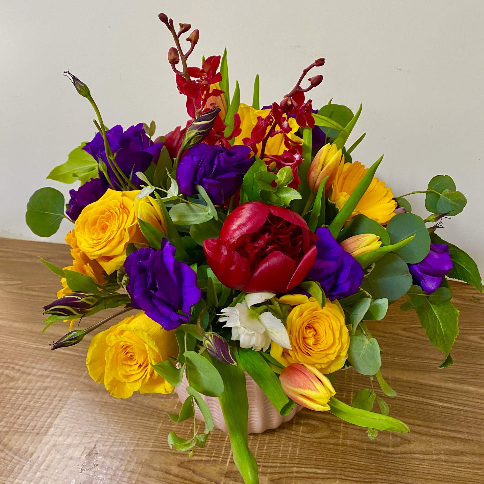 Bright arrangement of yellow roses, purple lisianthus, red peony and orange tulips in a pink vase