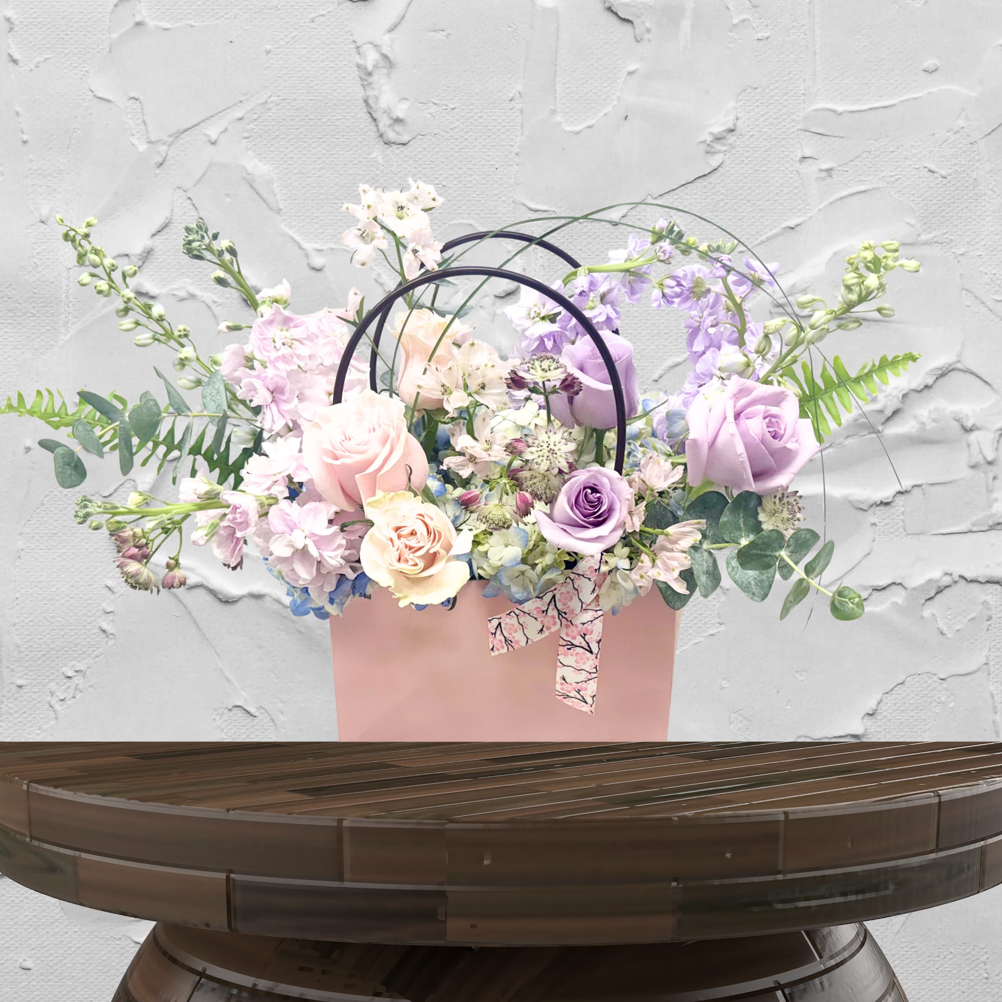 Pastel roses and lavender flowers arranged in a pink gift bag with ribbon handles.