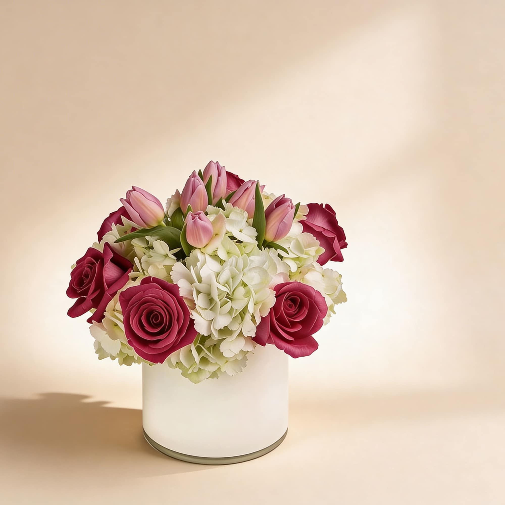 Low arrangement of pink roses, pink tulips, and white hydrangeas in a white cylinder vase