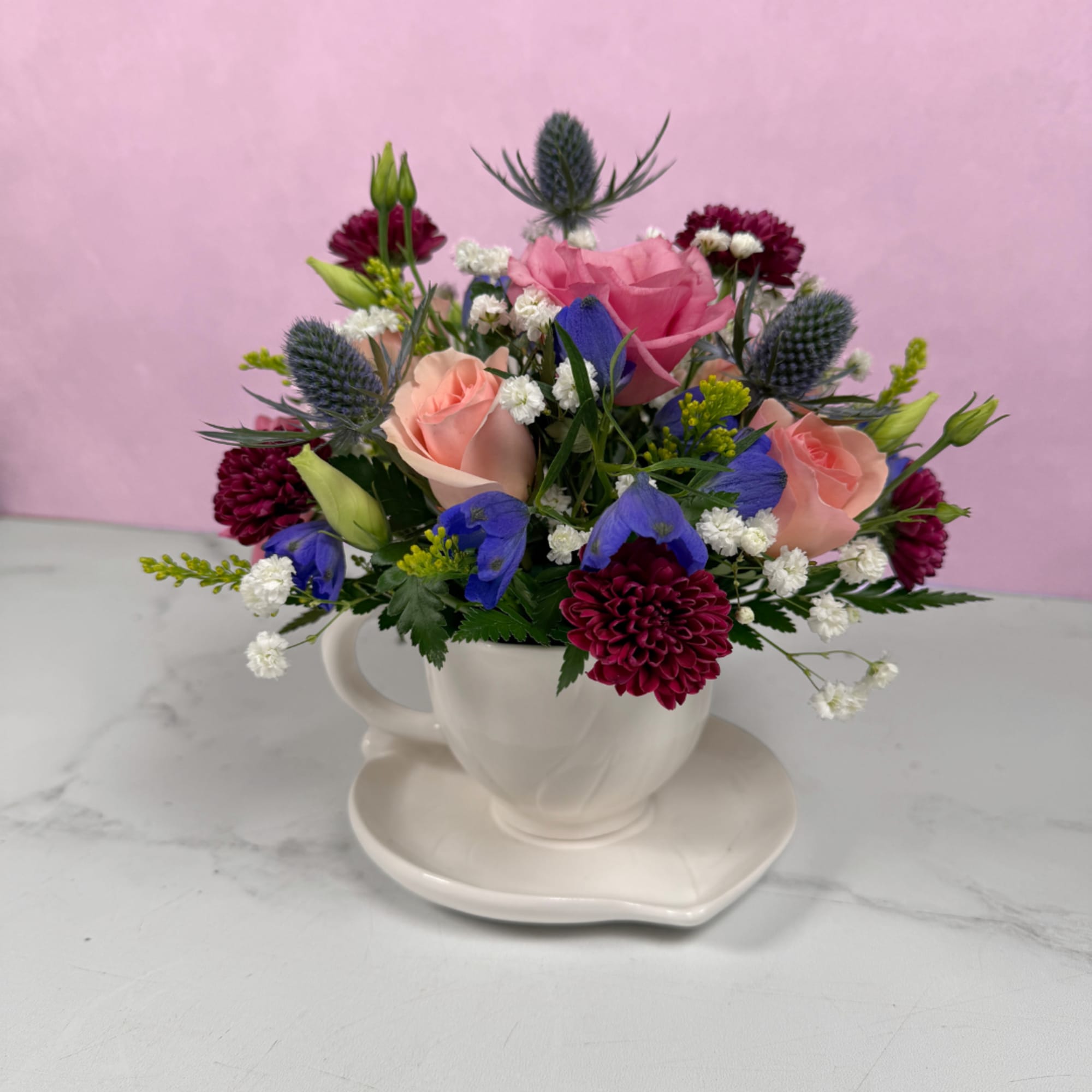 Soft blush roses and lively blue blooms spill from a white teacup