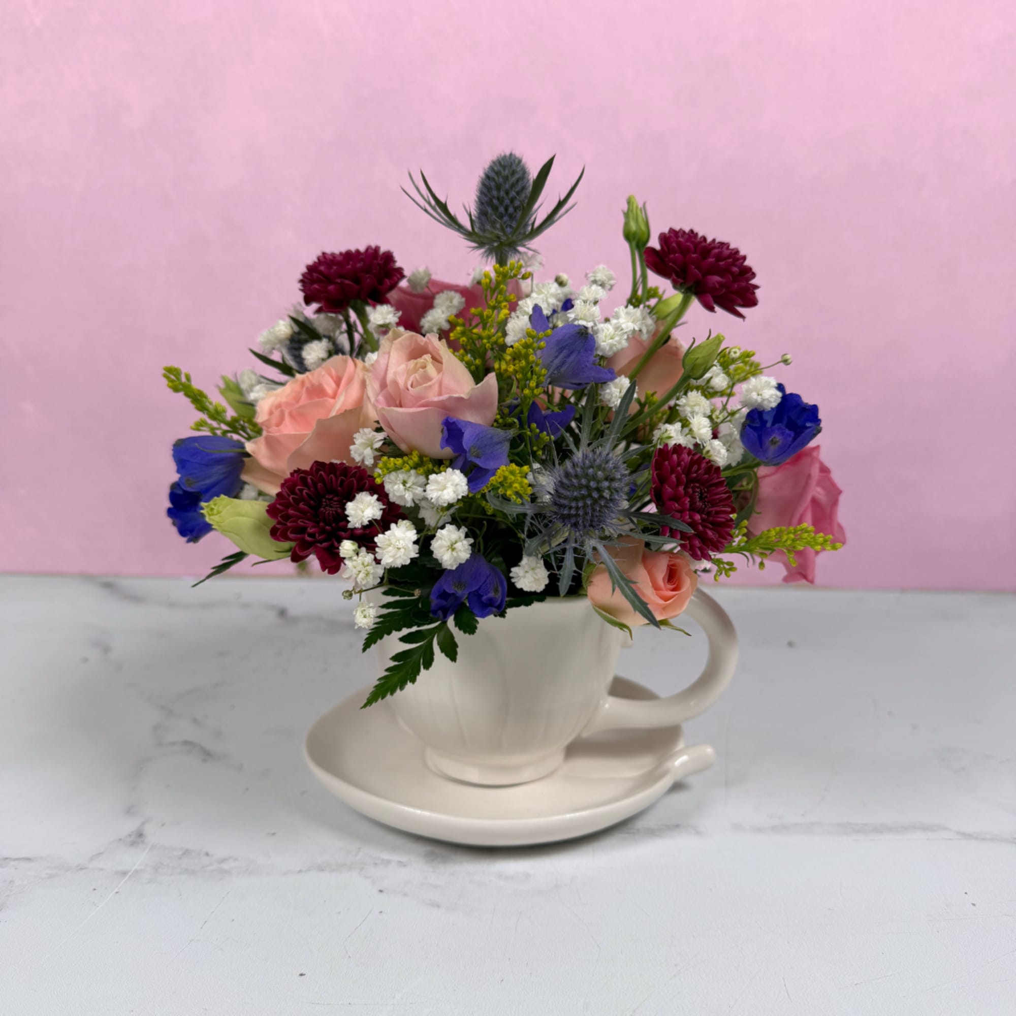 Soft blush roses and lively blue blooms spill from a white teacup