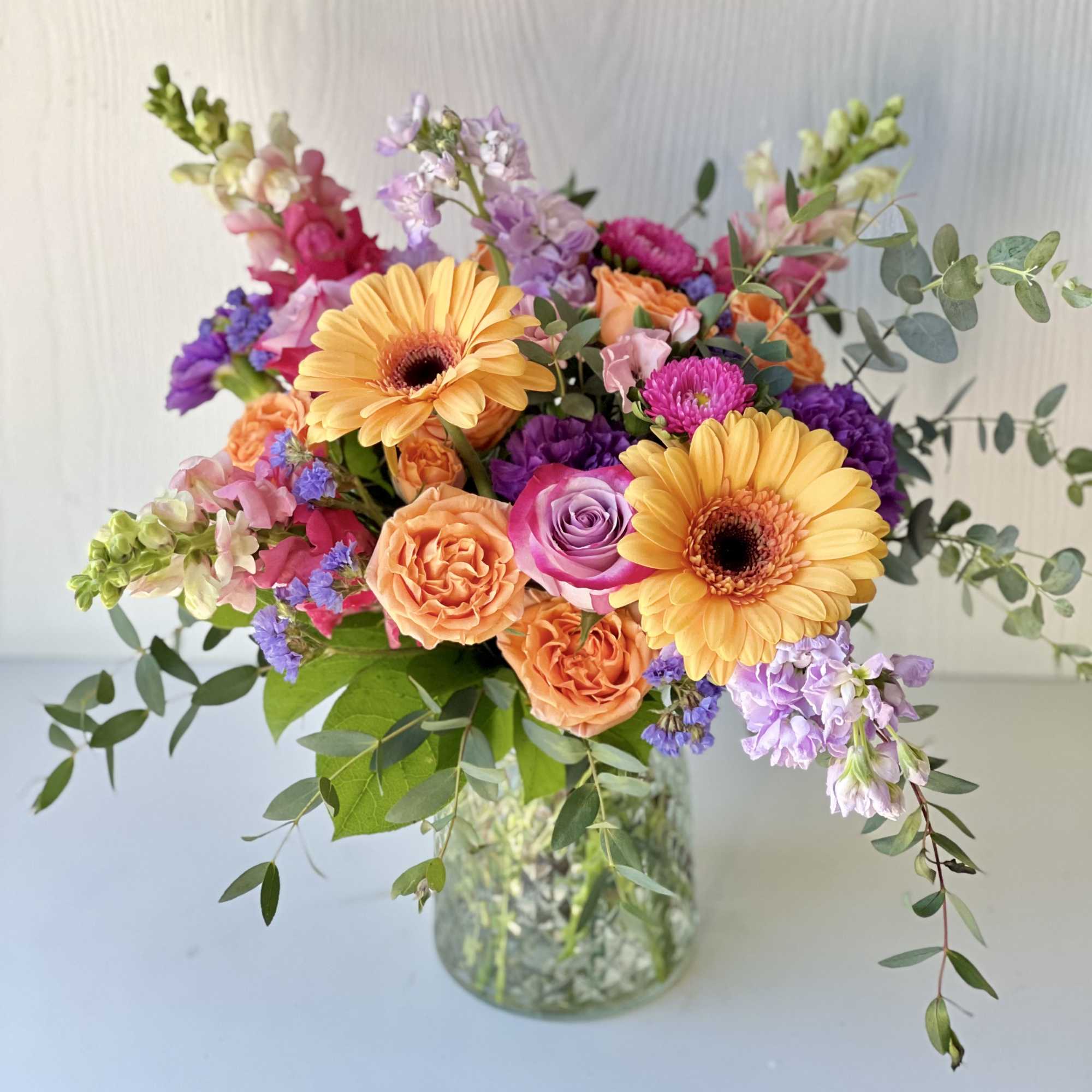 Mixed bouquet with orange gerberas, roses, and purple blooms in a glass vase
