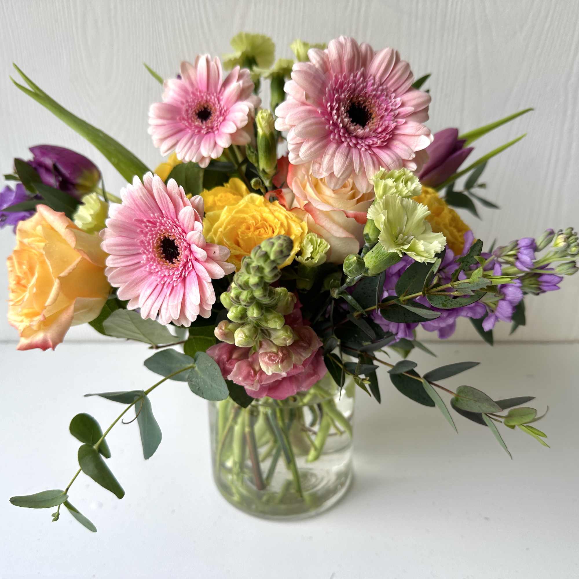 Mixed bouquet of pink gerbera daisies, roses, and purple flowers in a clear glass vase