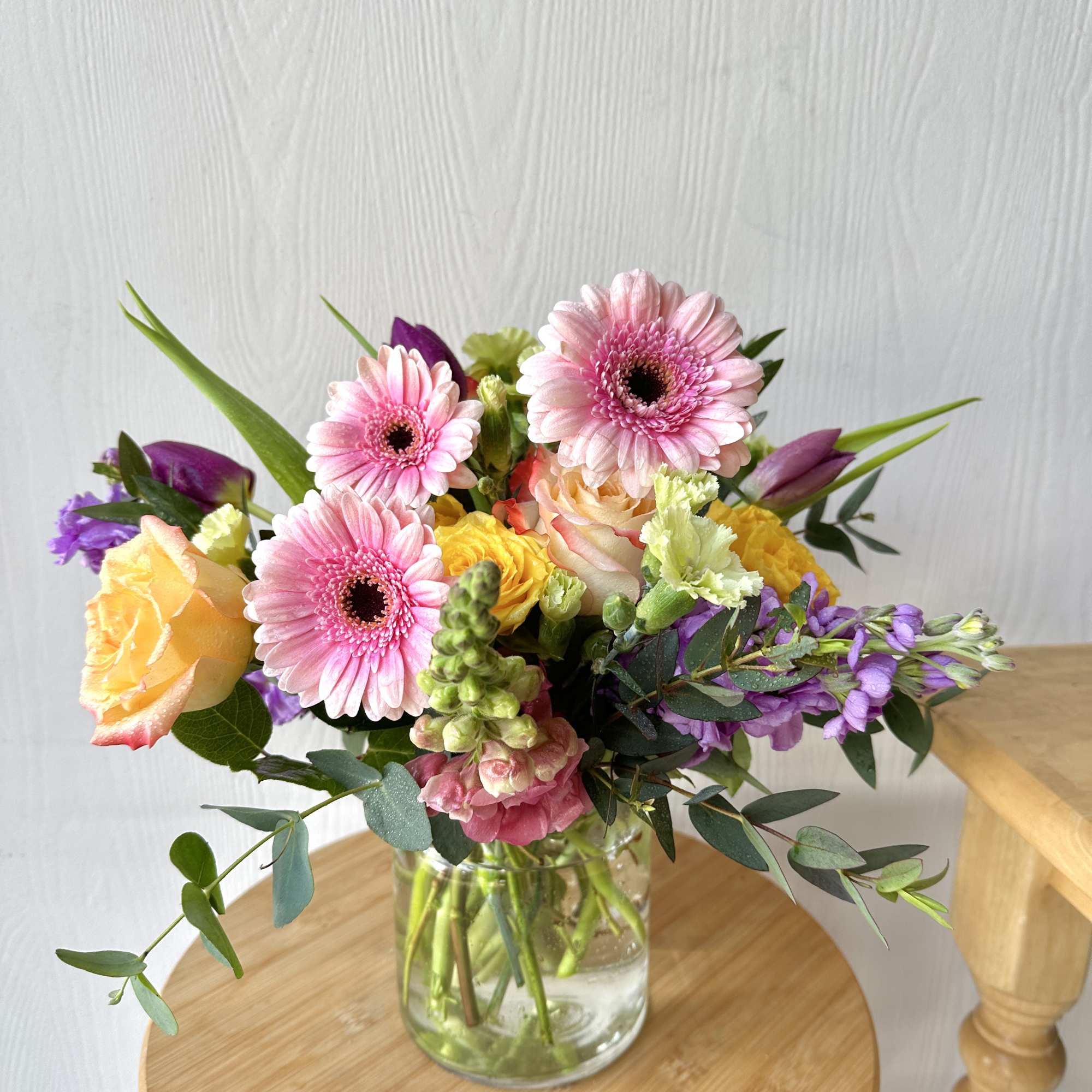 Mixed bouquet of pink gerbera daisies, roses, and purple blooms in a glass vase