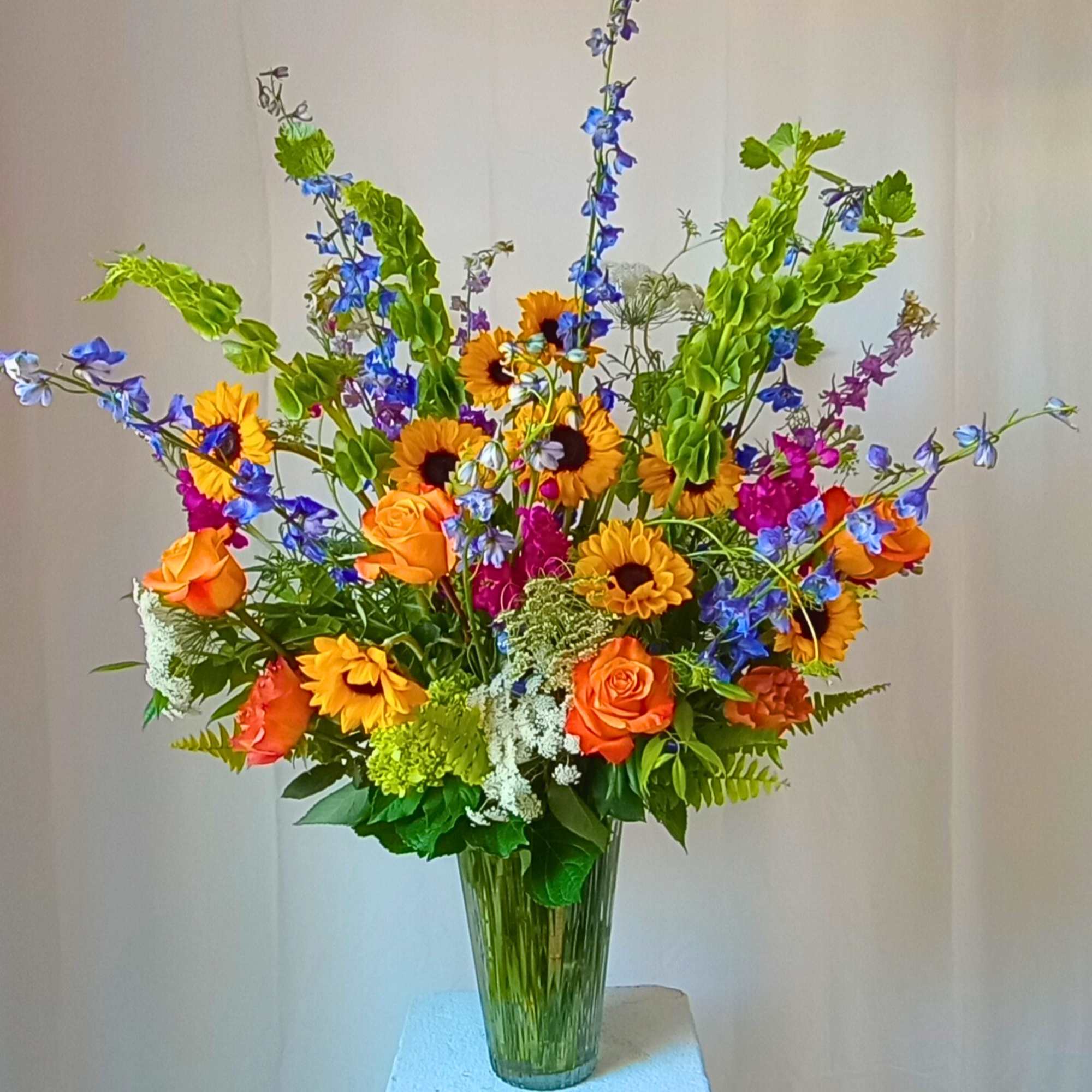 Tall mixed bouquet with orange roses, sunflowers, and blue flowers in a glass vase