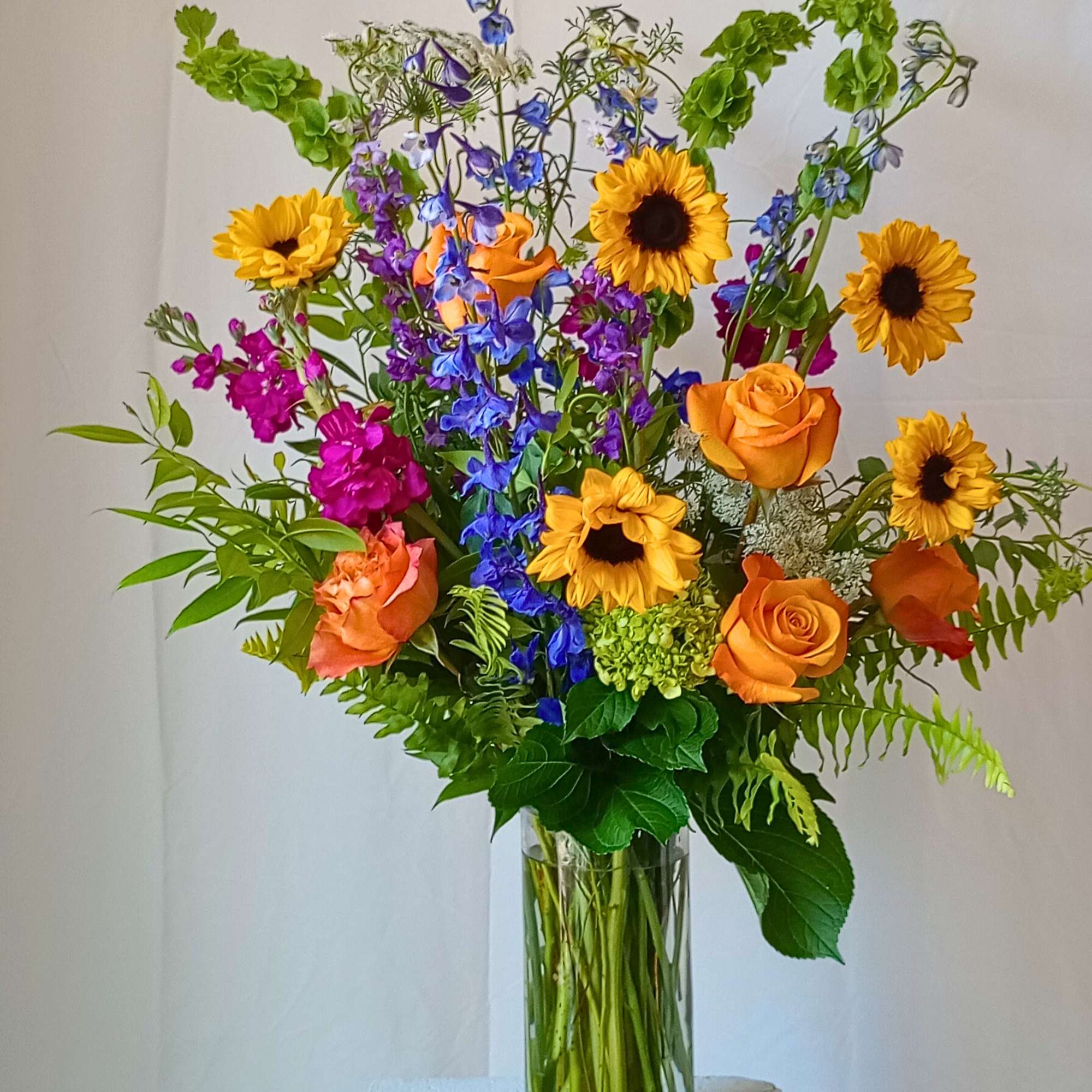 Tall mixed bouquet with sunflowers, orange roses, and blue flowers in a glass vase