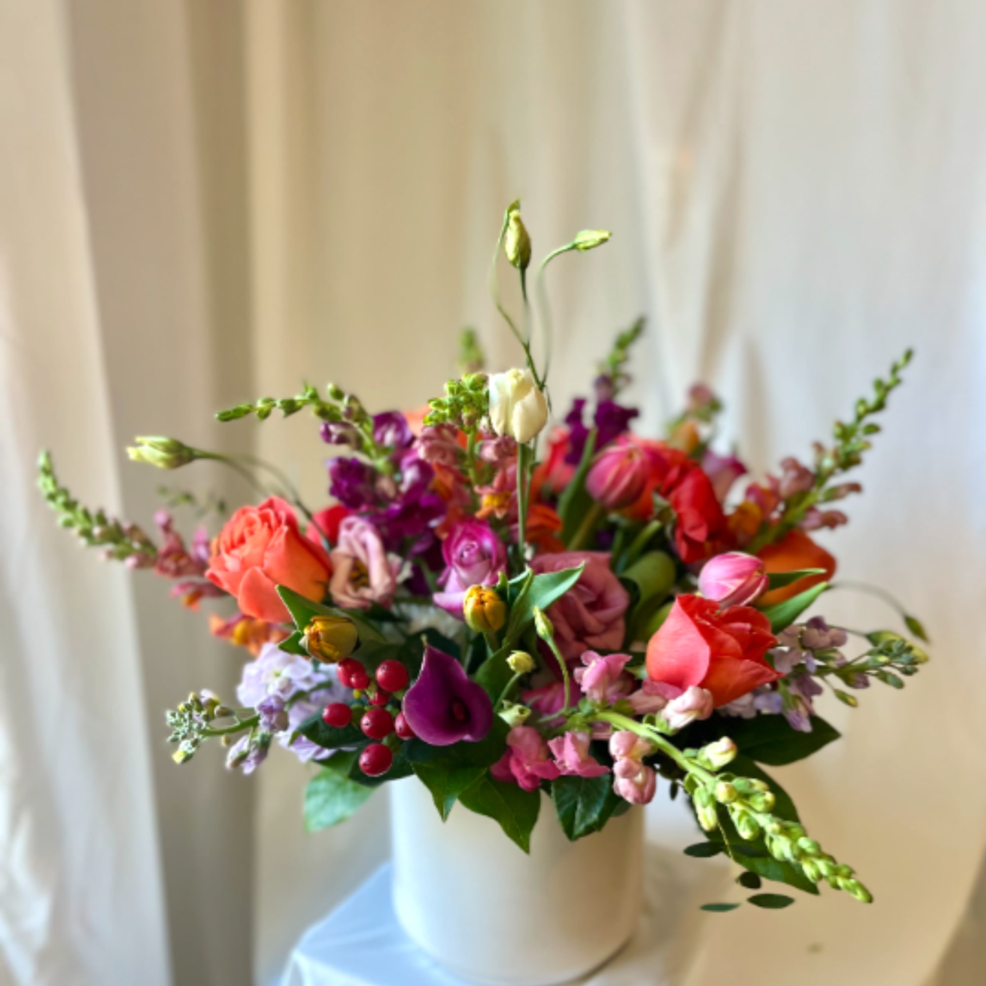 Colorful arrangement of orange roses, pink and purple blooms in a white vase on a draped pedestal