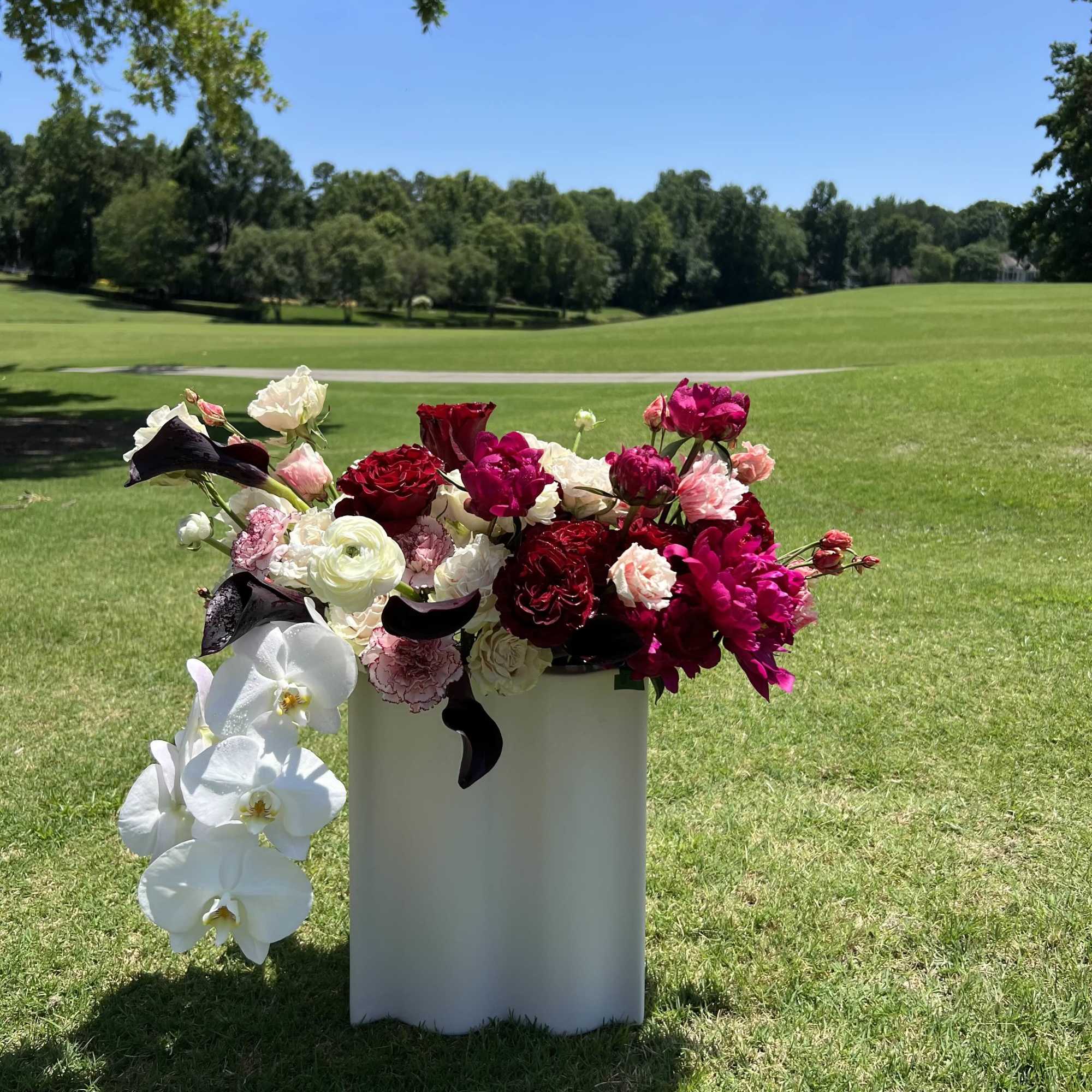 Mixed pink, red, and white flowers arranged in a white pedestal vase outdoors.