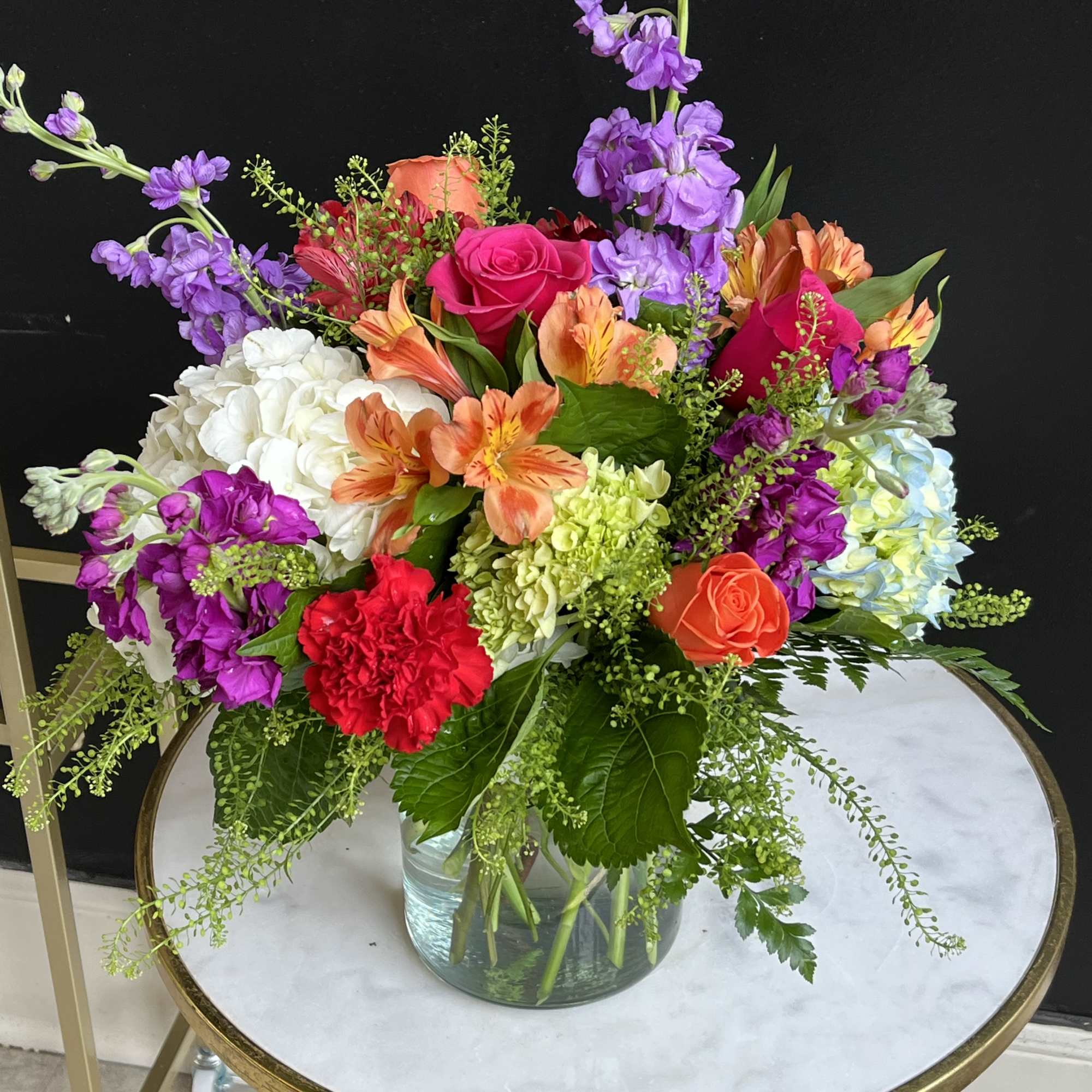 Colorful mixed bouquet in a clear glass vase with roses, hydrangeas, and purple blooms