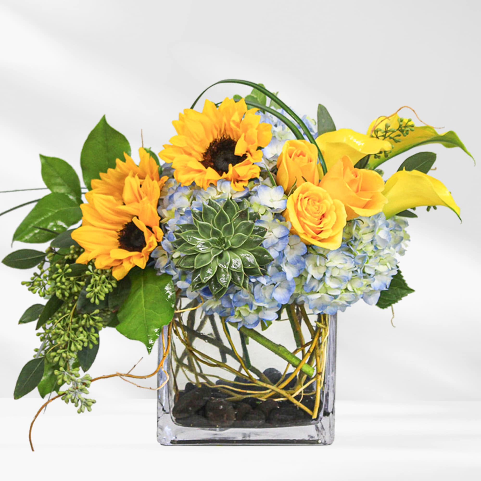 Sunflowers, yellow roses, blue hydrangeas, and calla lilies in a square glass vase
