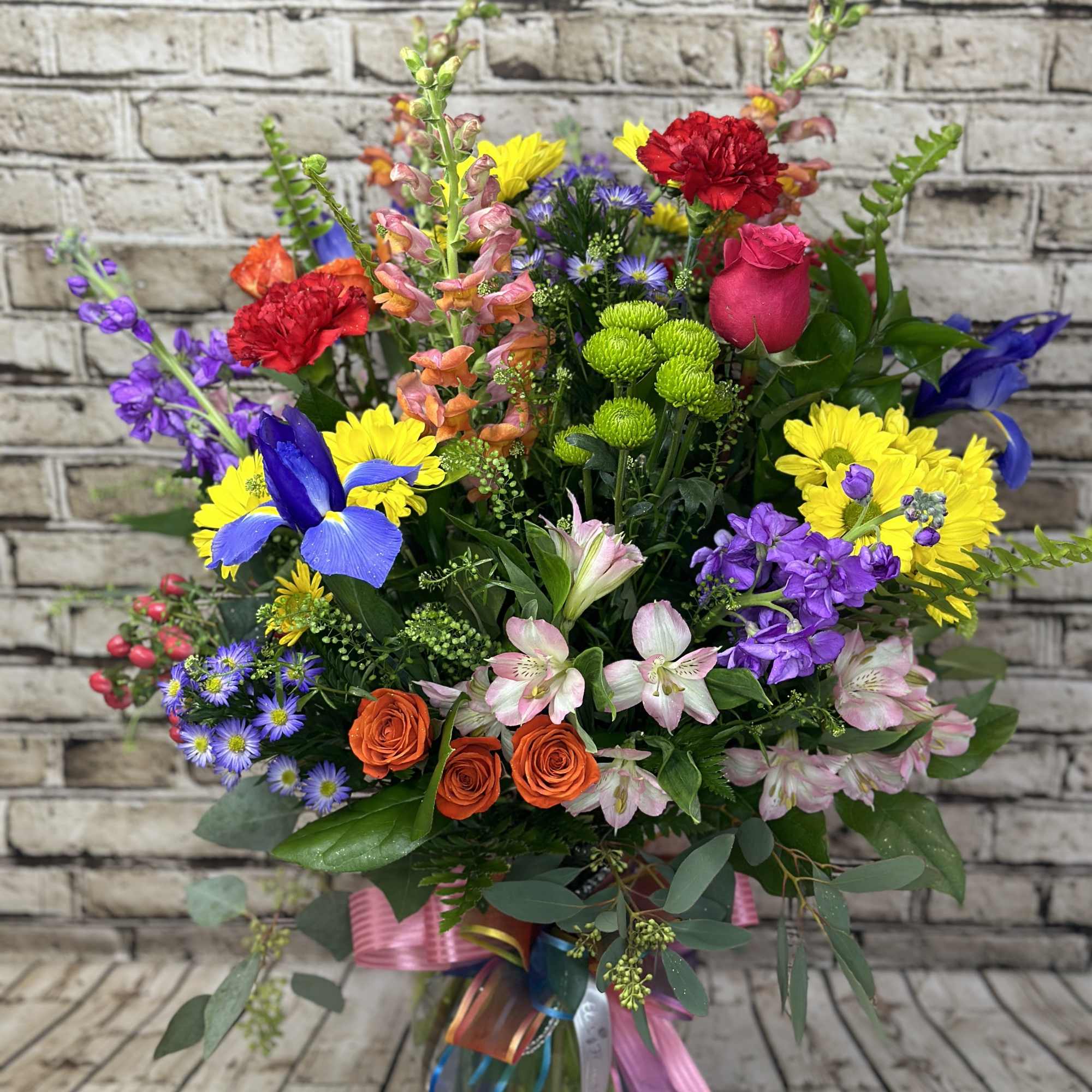 Tall mixed bouquet of bright roses, daisies, irises, asters and more in a clear vase with colorful ribbons.
