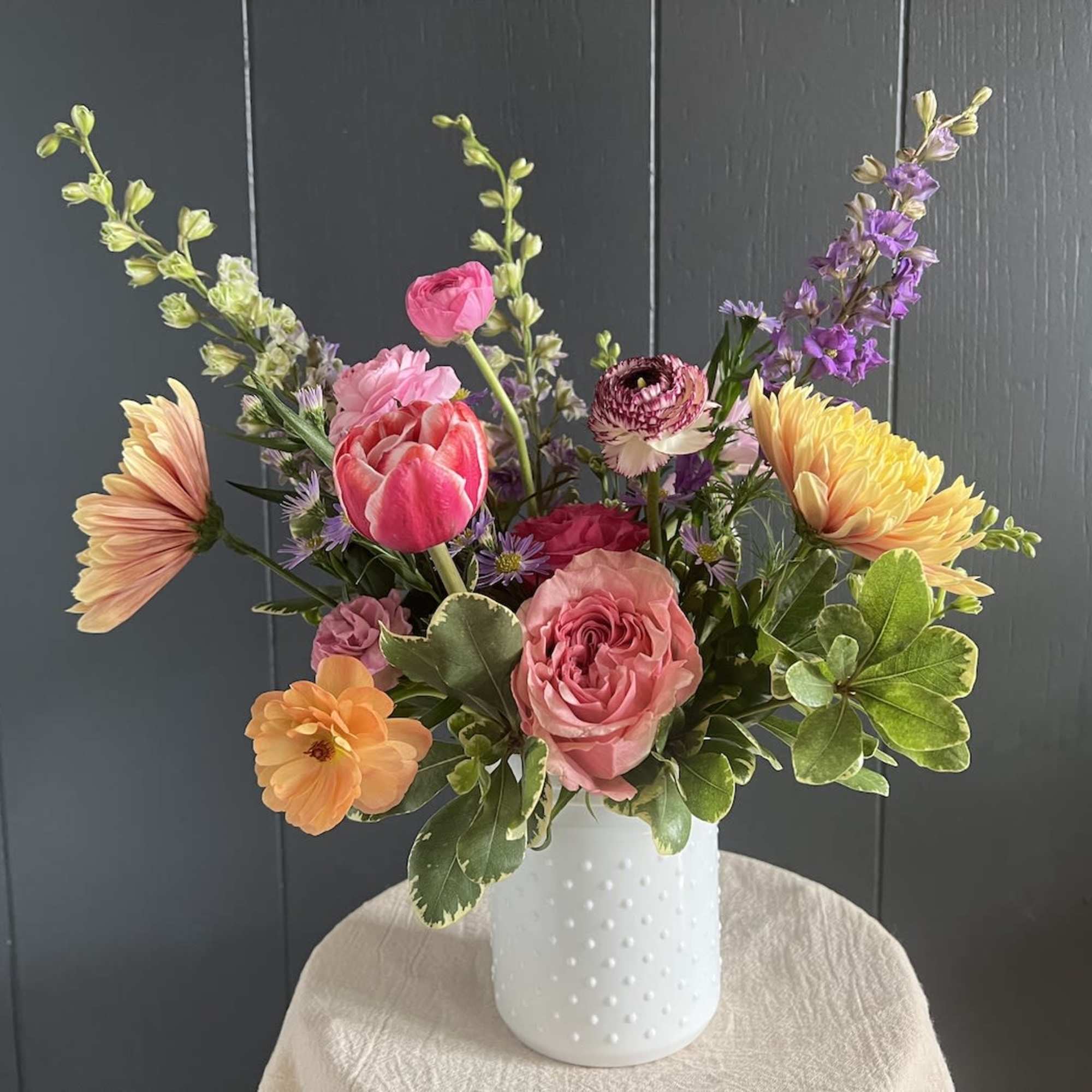 Mixed bouquet of pink, peach, yellow, and purple flowers in a white vase