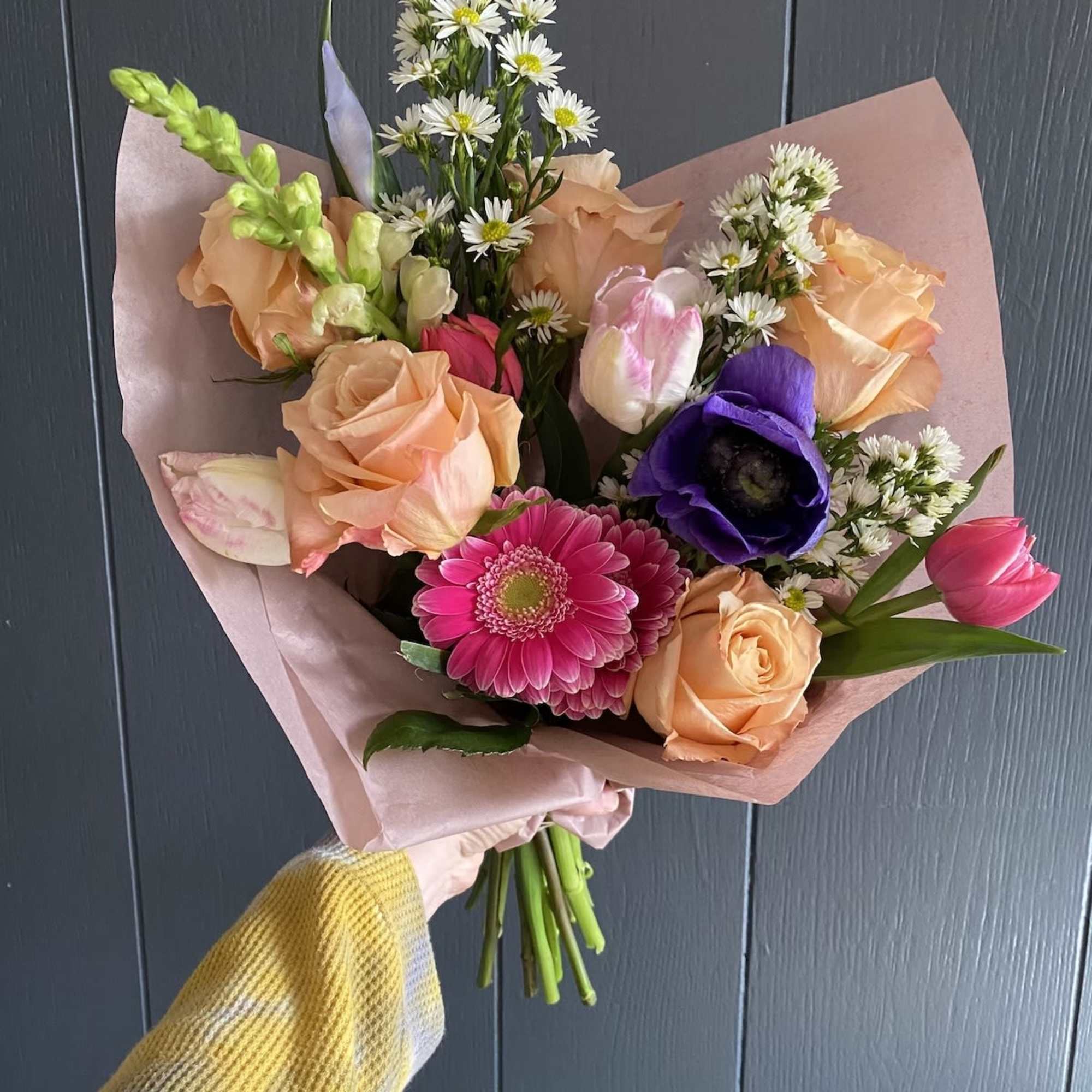 Handheld bouquet of peach roses, pink gerbera daisies, and purple blooms