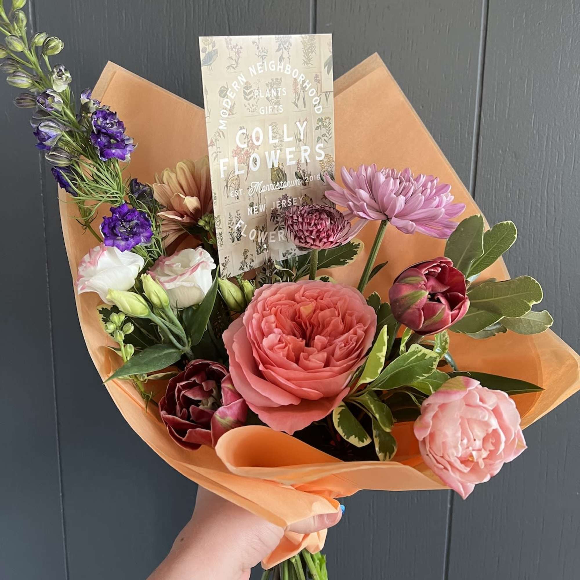 Handheld bouquet of mixed pink, purple, and peach flowers wrapped in orange paper