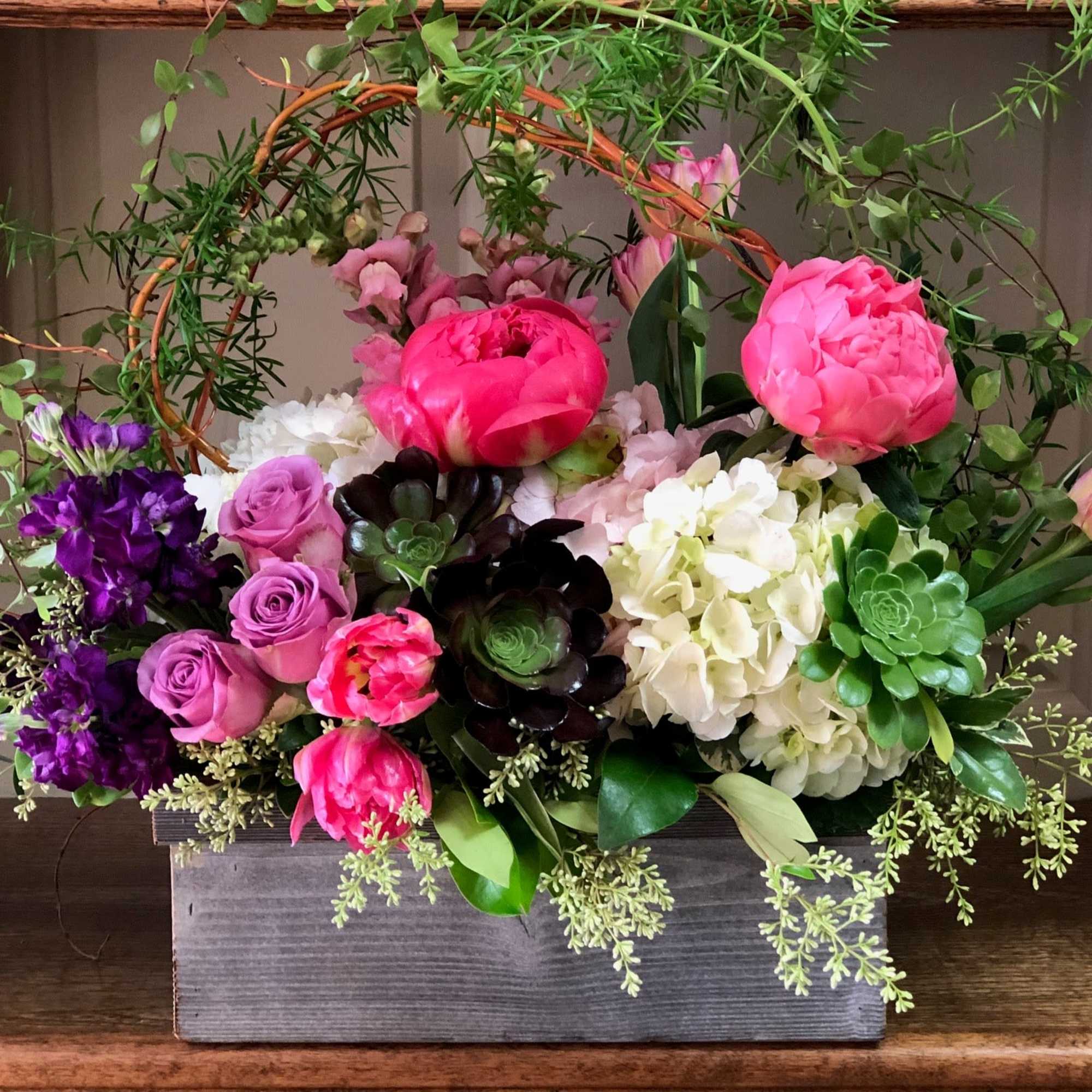 Arrangement of pink peonies, lavender roses, white hydrangeas, tulips, and succulents in a gray wooden box.