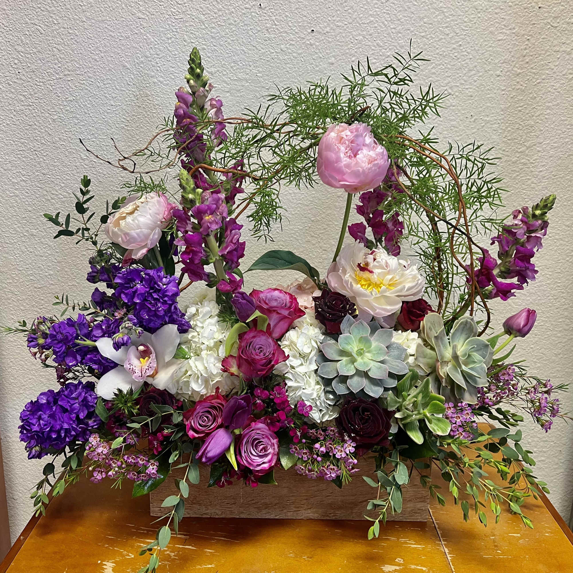 Lush rectangular arrangement with purple and pink blooms, white hydrangeas, and succulents in a wooden box.