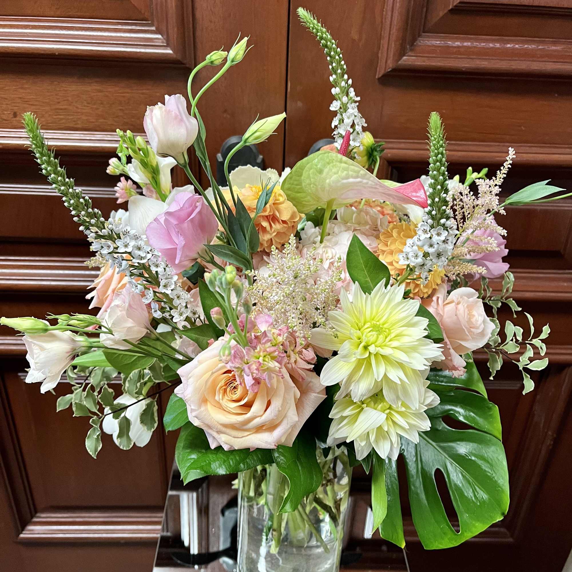 Tall vase arrangement of peach roses, white dahlias, and pink lisianthus with a large green leaf.