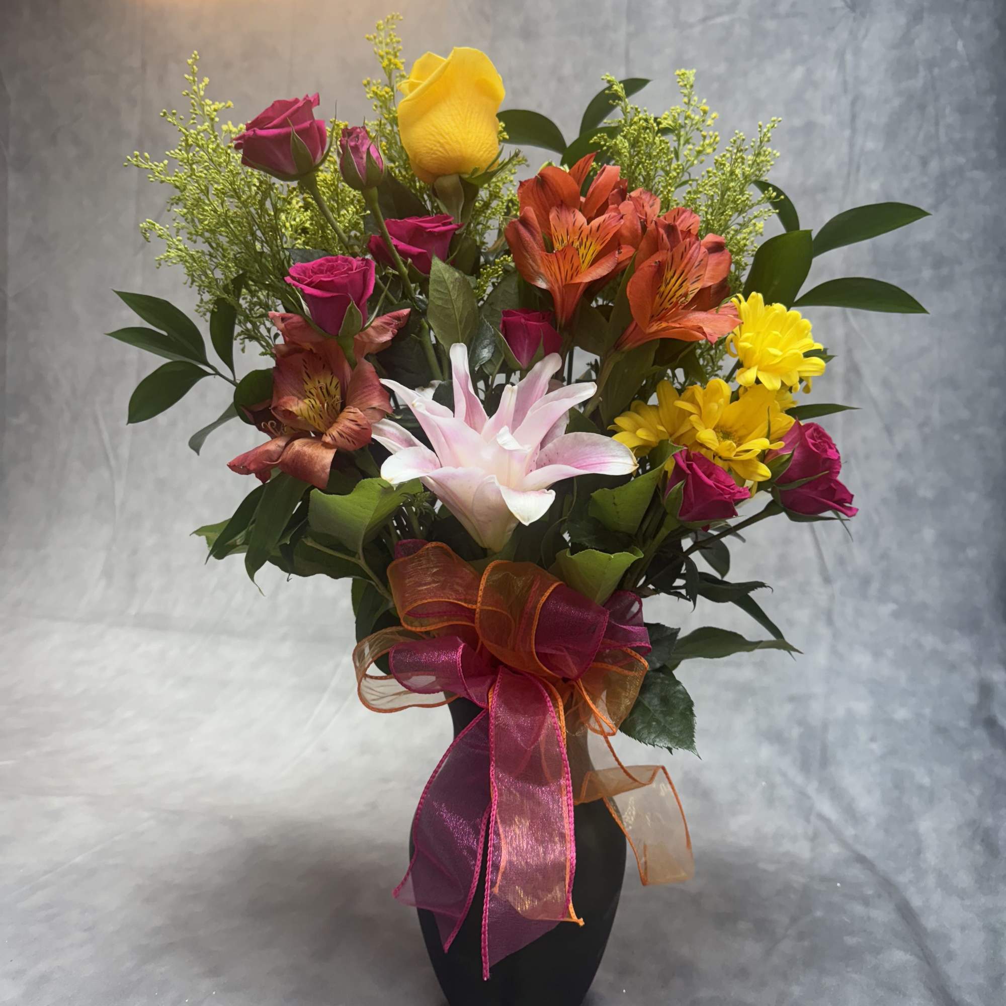 Mixed bouquet of roses, lilies, and daisies in a black vase with ribbon