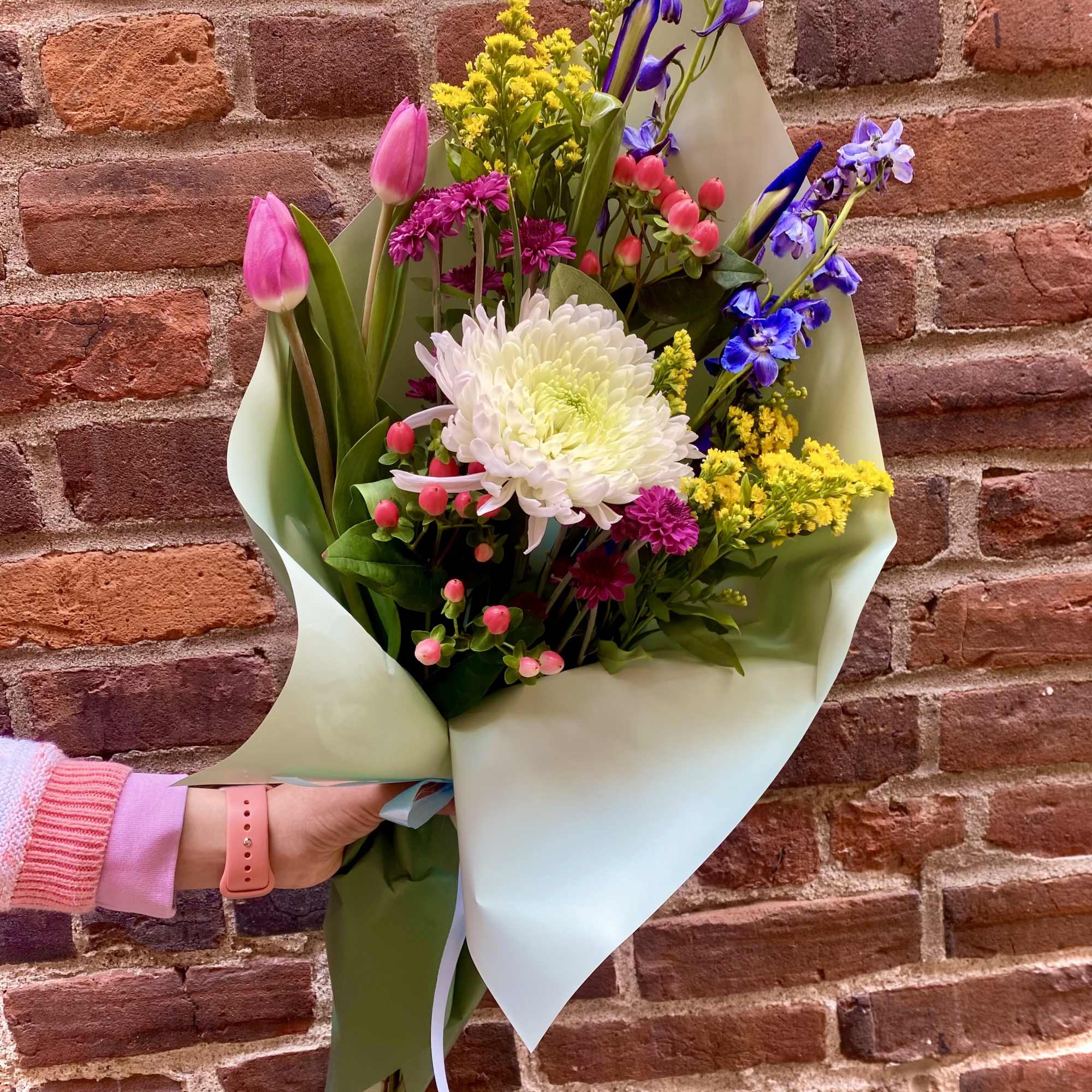 Handheld bouquet of pink tulips, white chrysanthemum, and purple flowers in paper wrap