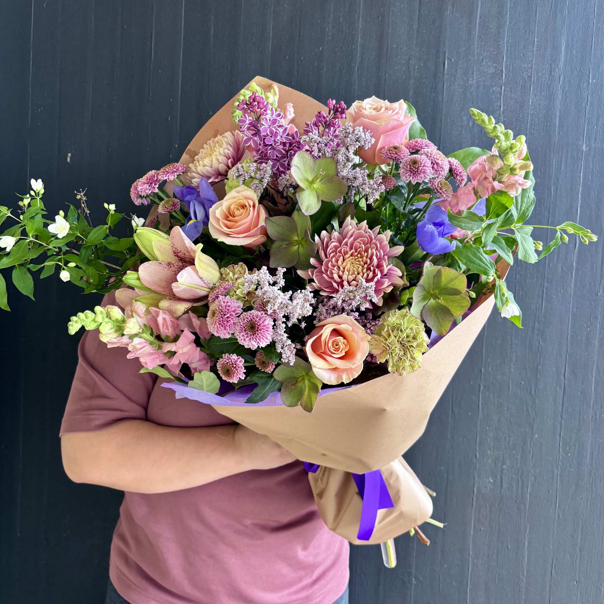 Large hand-tied bouquet of pink and peach flowers wrapped in kraft paper with purple ribbon