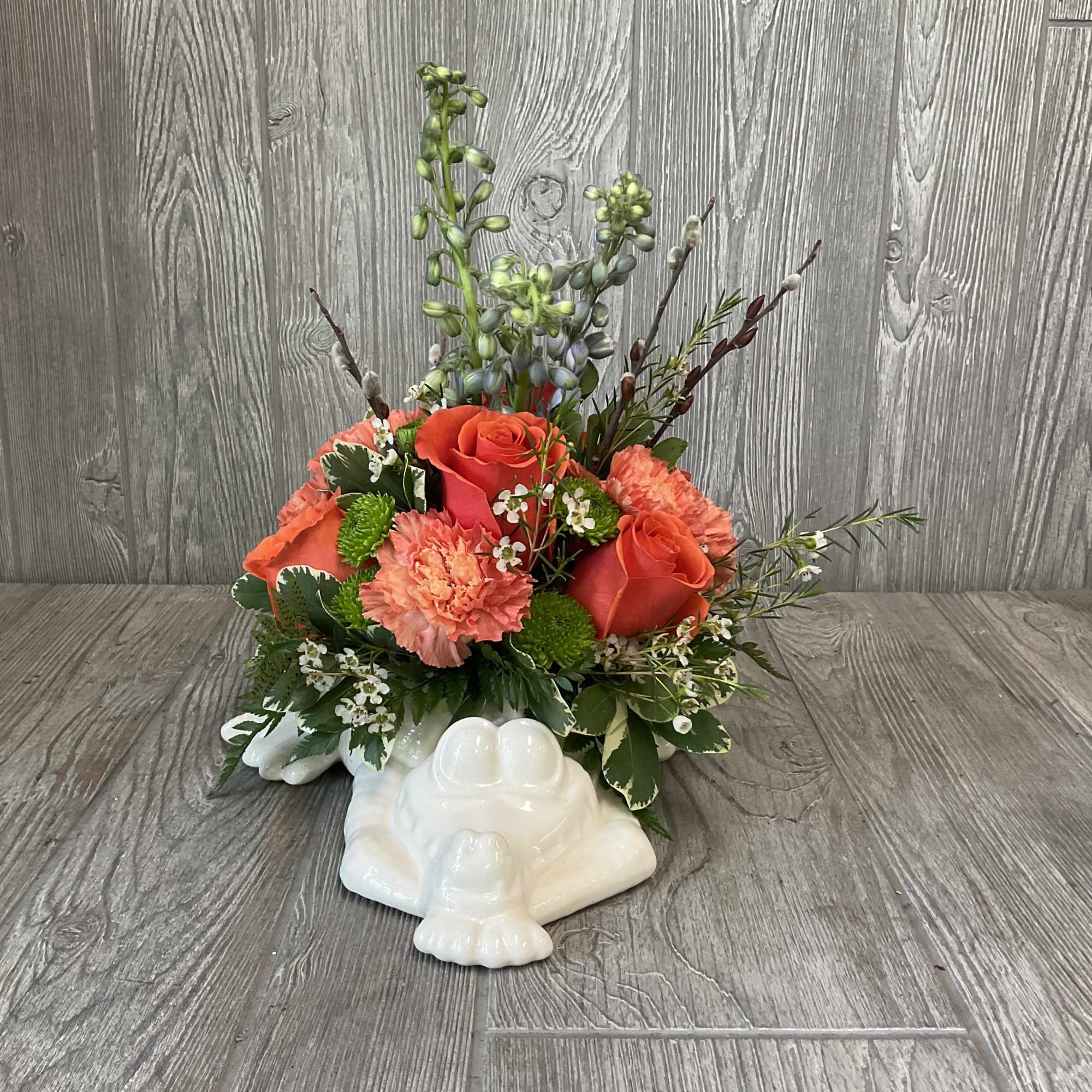 Compact arrangement of orange roses and carnations in a white ceramic frog container