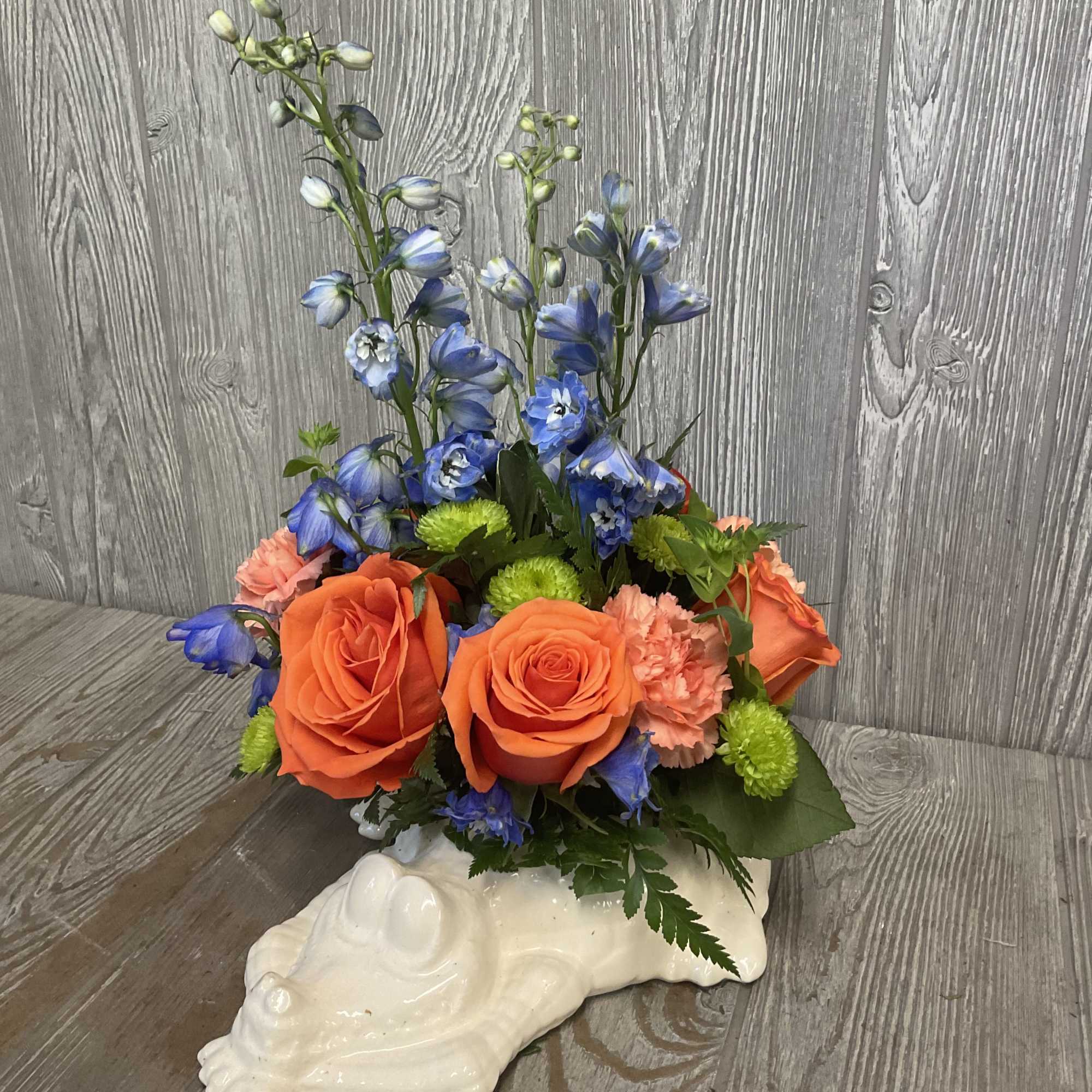 Flower arrangement with orange roses, blue delphinium, and pink carnations in a white ceramic alligator container