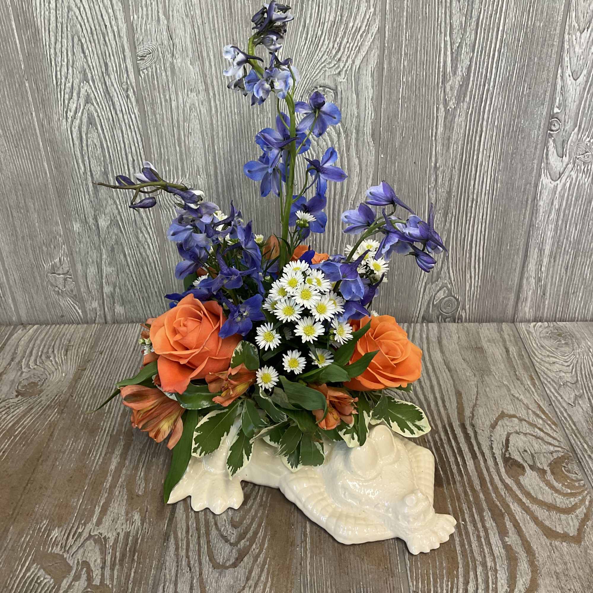 Orange roses, blue delphinium, and white daisies in a white ceramic turtle container