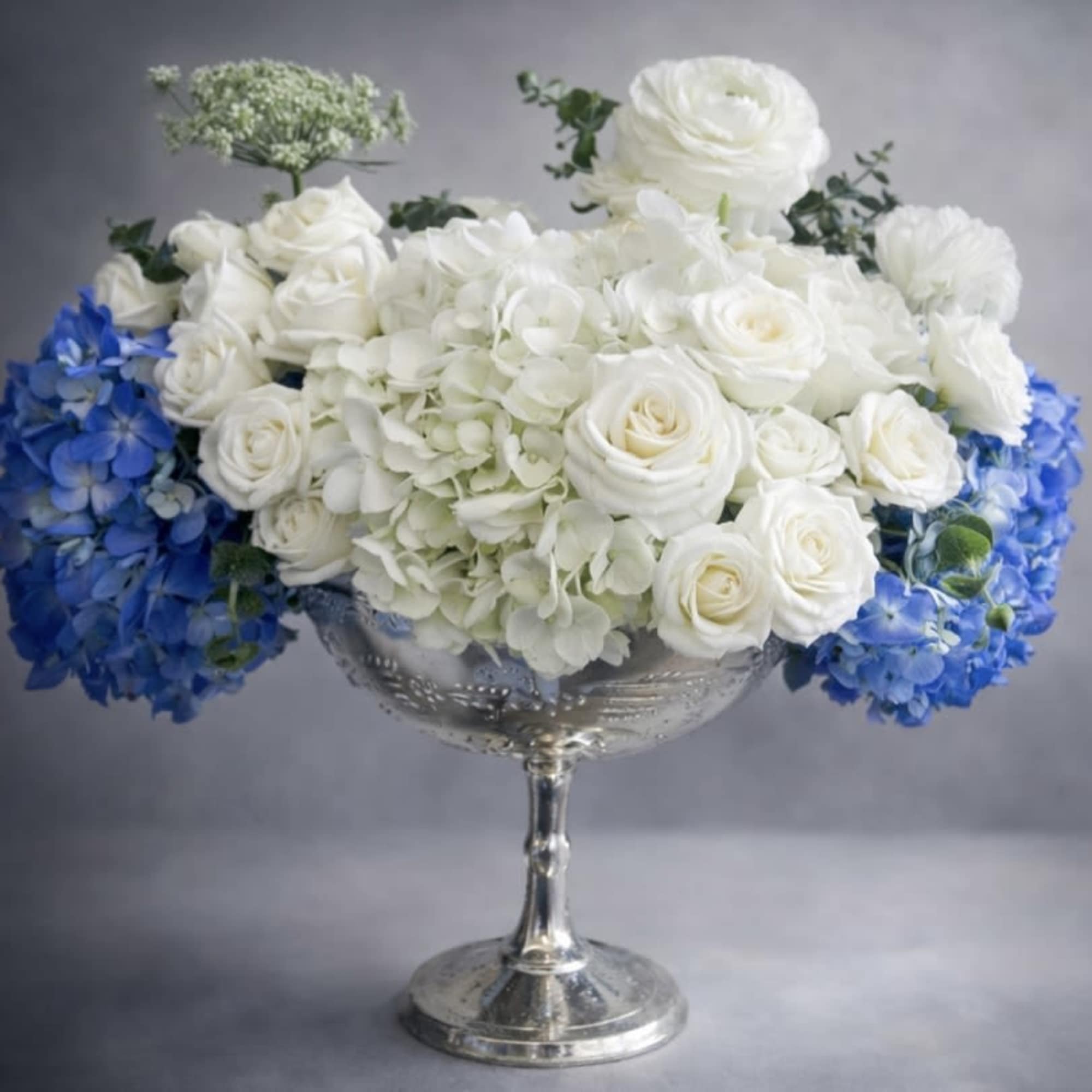 White roses and hydrangeas arranged in a silver pedestal bowl with blue flowers at the sides