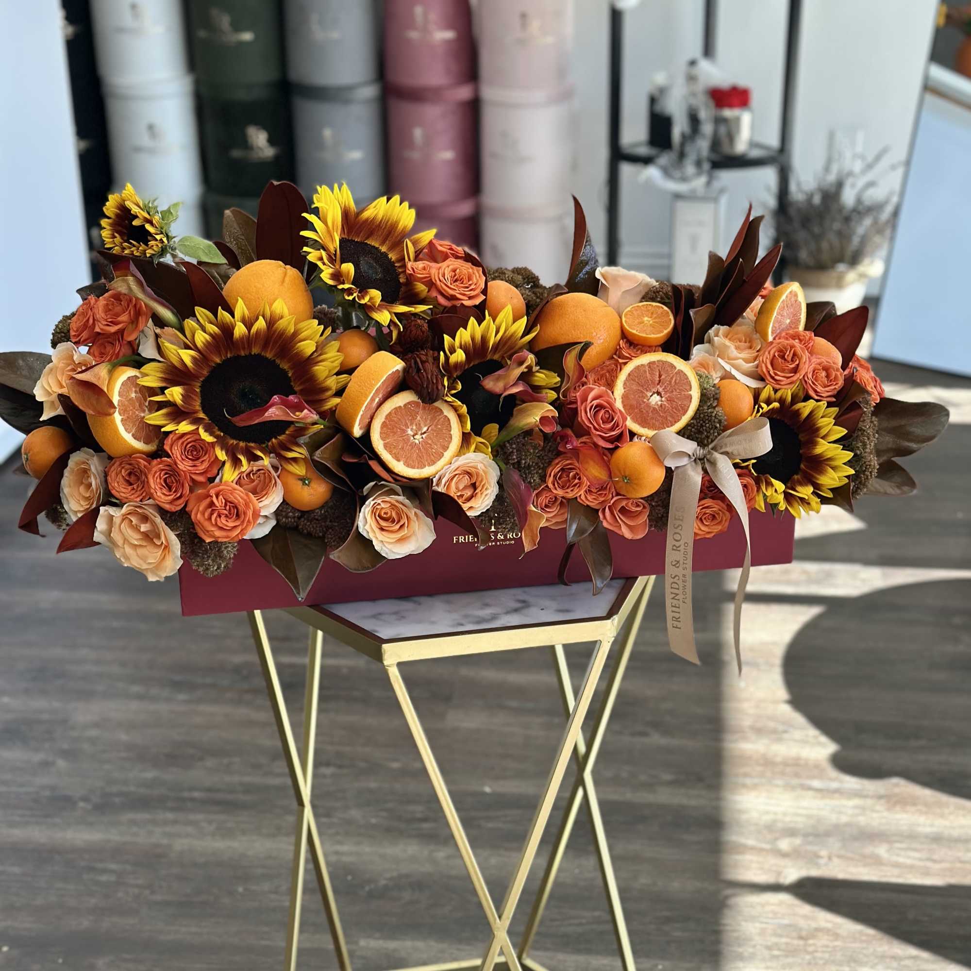 Low floral arrangement with sunflowers, orange roses, and sliced citrus in a burgundy box.
