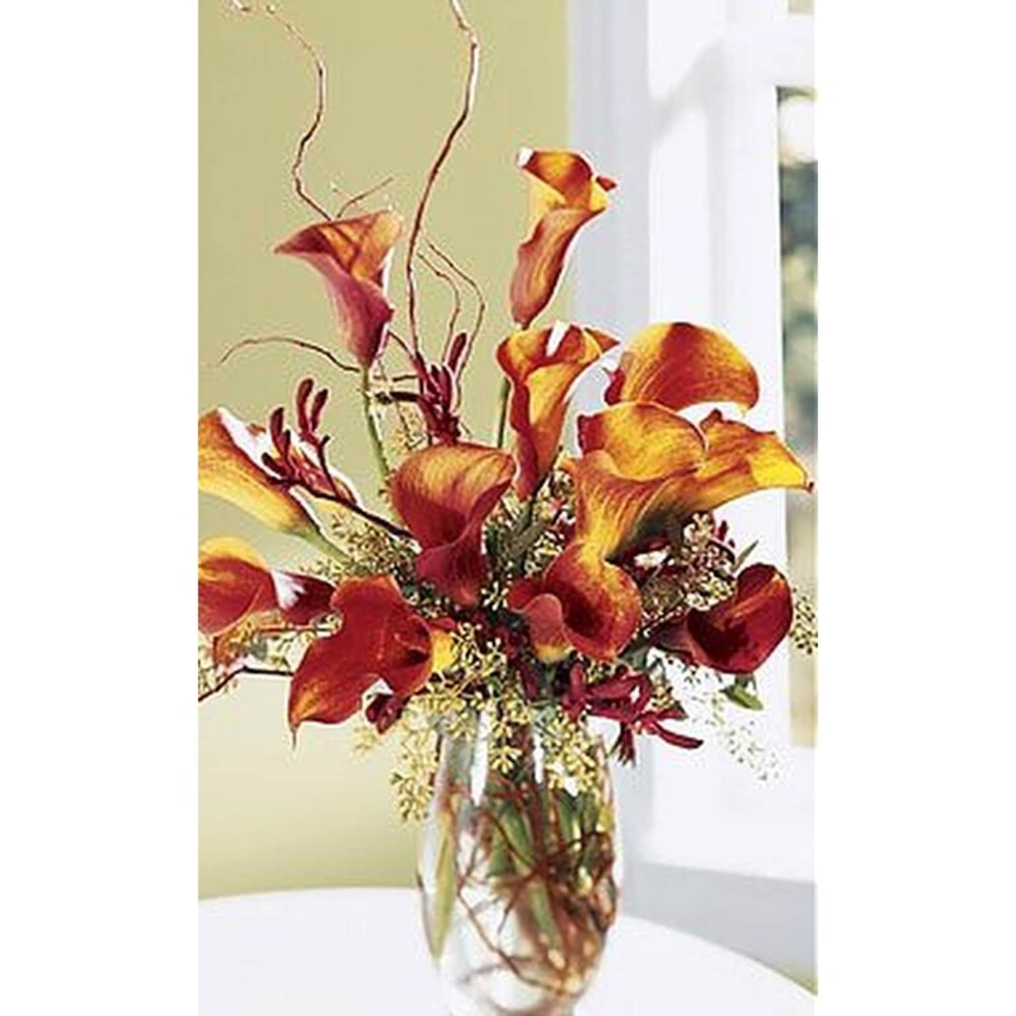 Tall arrangement of orange and red calla lilies in a clear glass vase with curly branches.