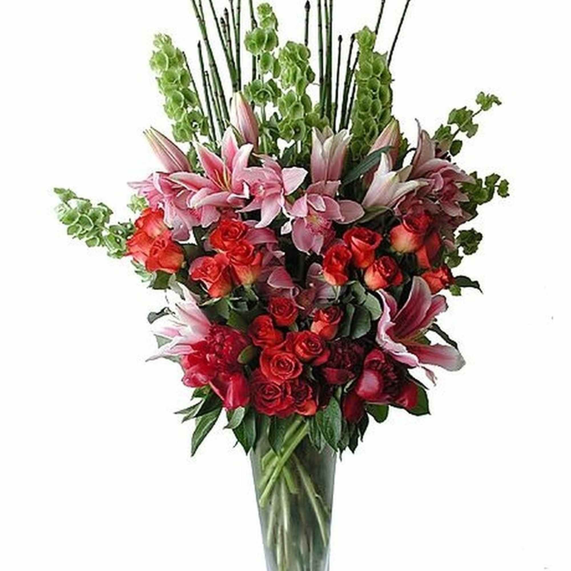 Tall arrangement of pink lilies, red roses, and green bells of Ireland in a clear glass vase.