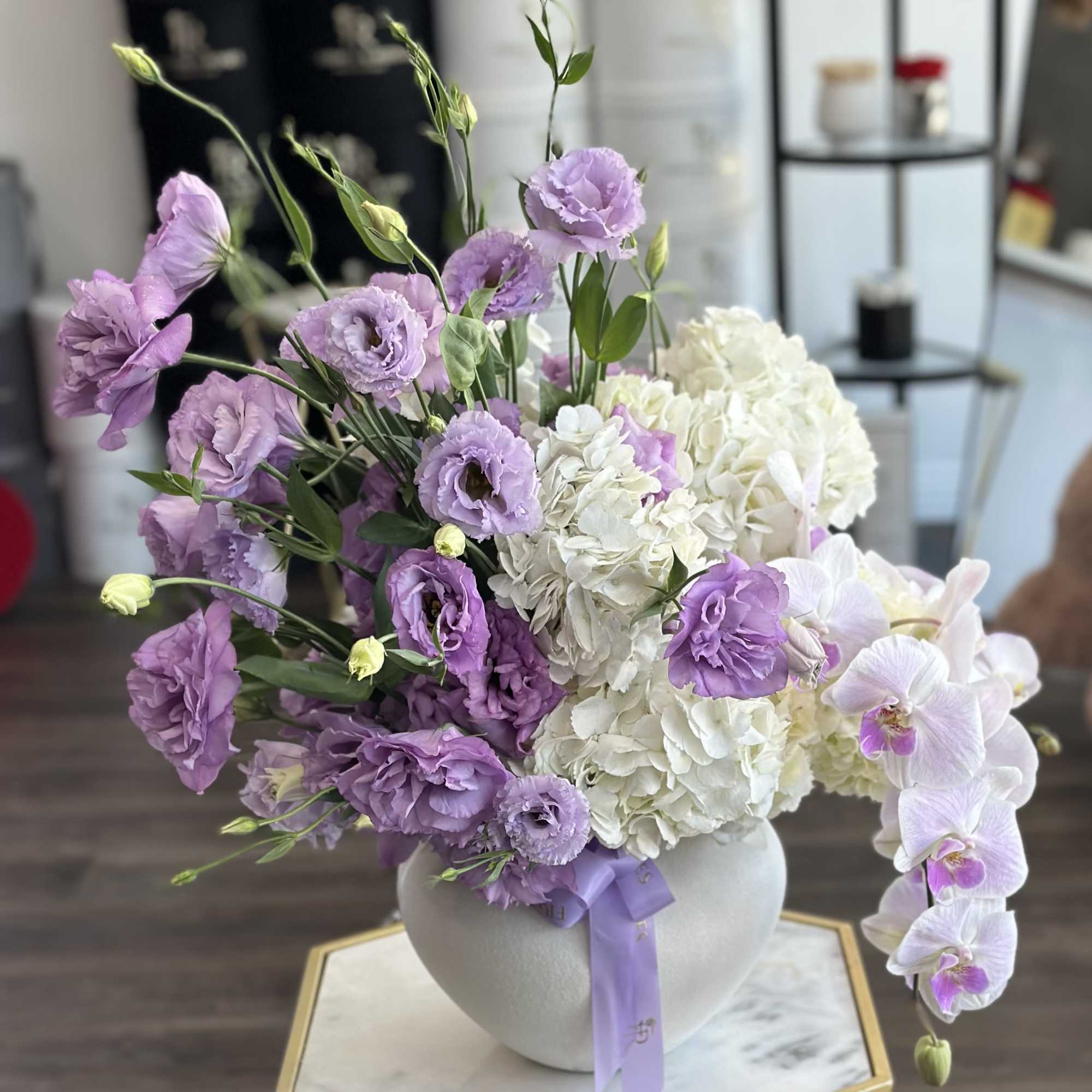 Lavender and white flowers arranged in a round vase with a purple ribbon