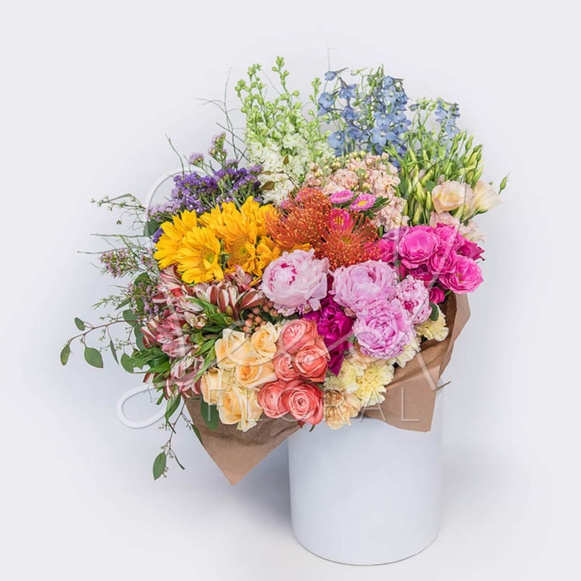 Large mixed-color bouquet with sunflowers, roses, peonies, and protea wrapped in kraft paper in a white cylinder.