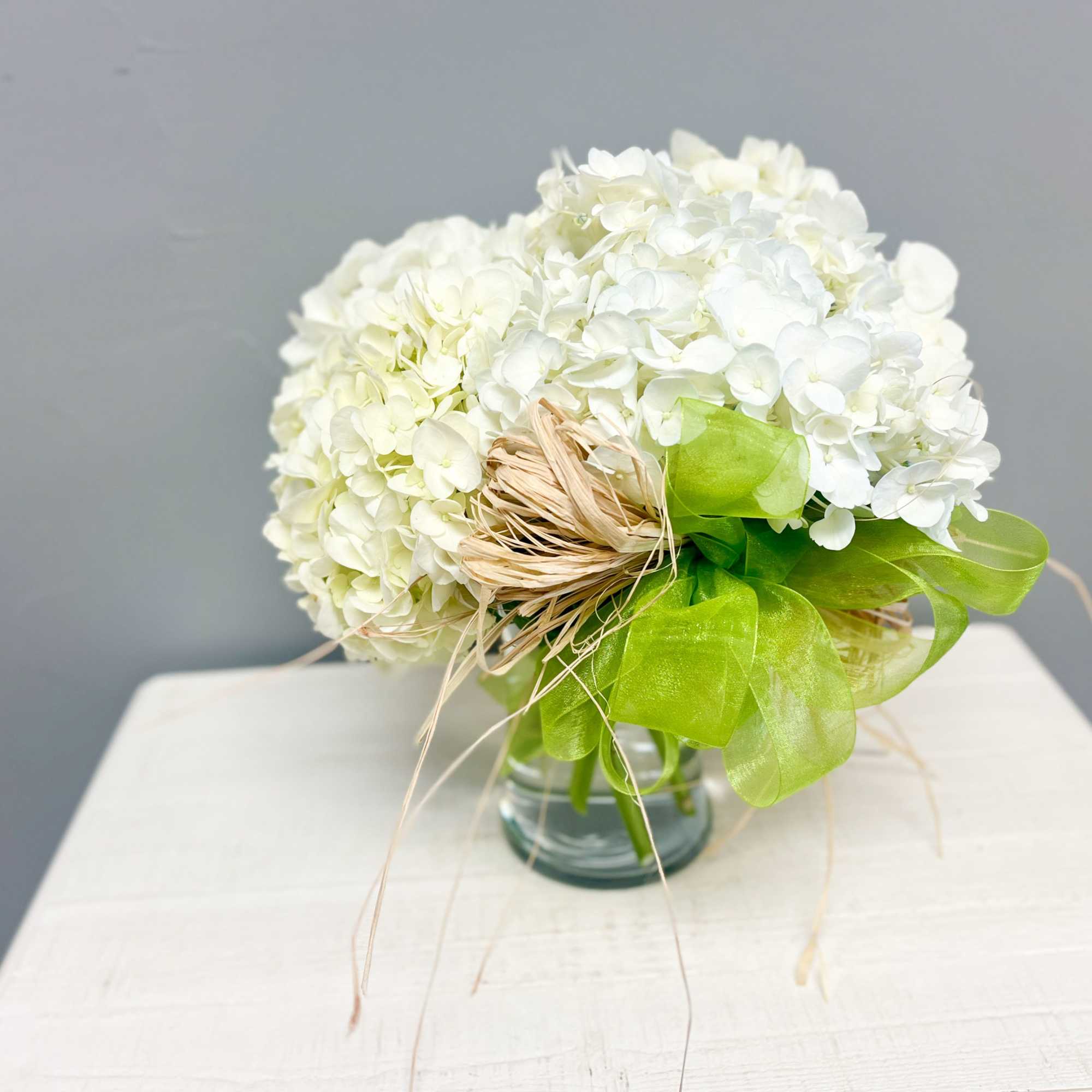 Round arrangement of white hydrangeas in a clear glass vase with green ribbon and raffia accents
