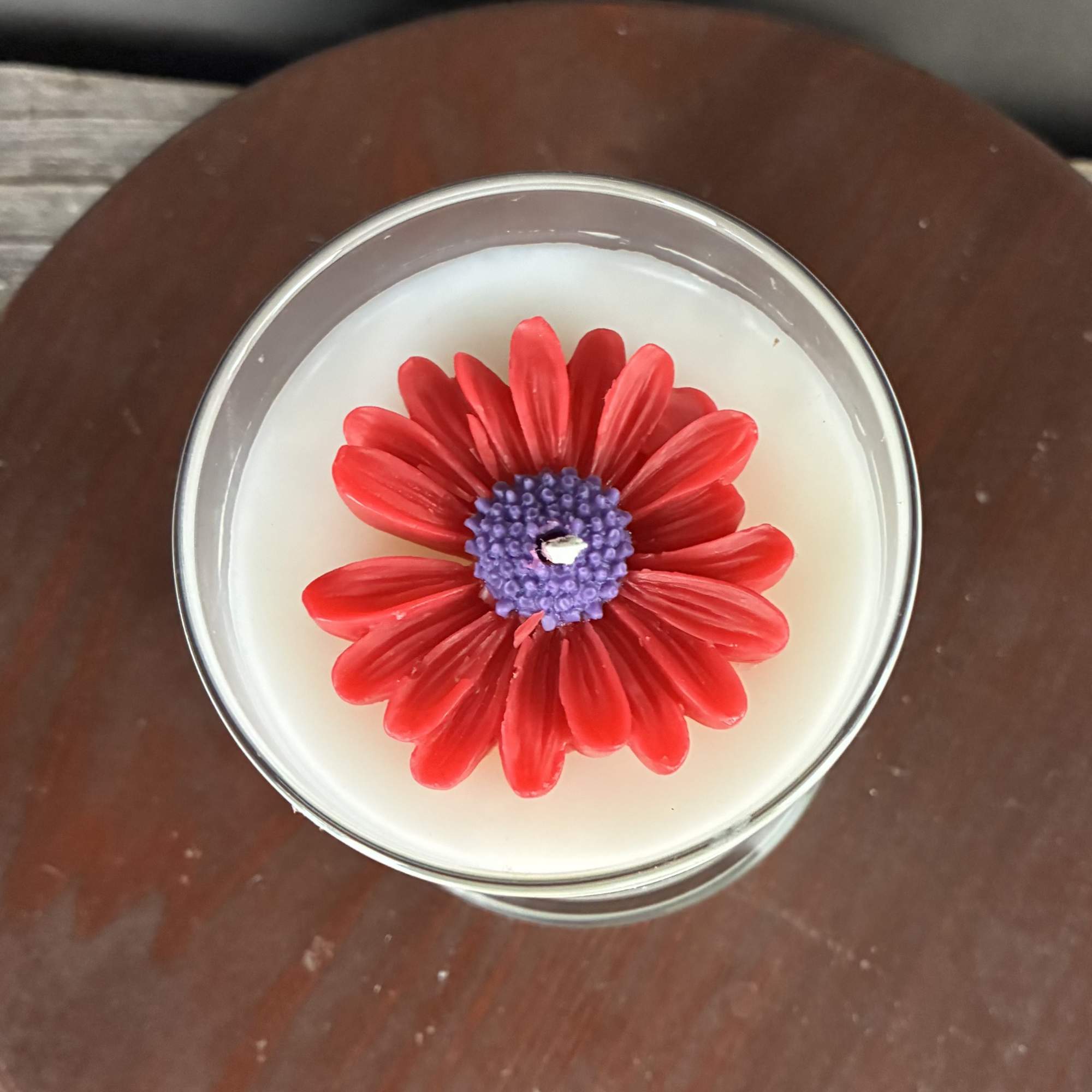 White candle in a glass jar topped with a red flower-shaped wax design with purple center.