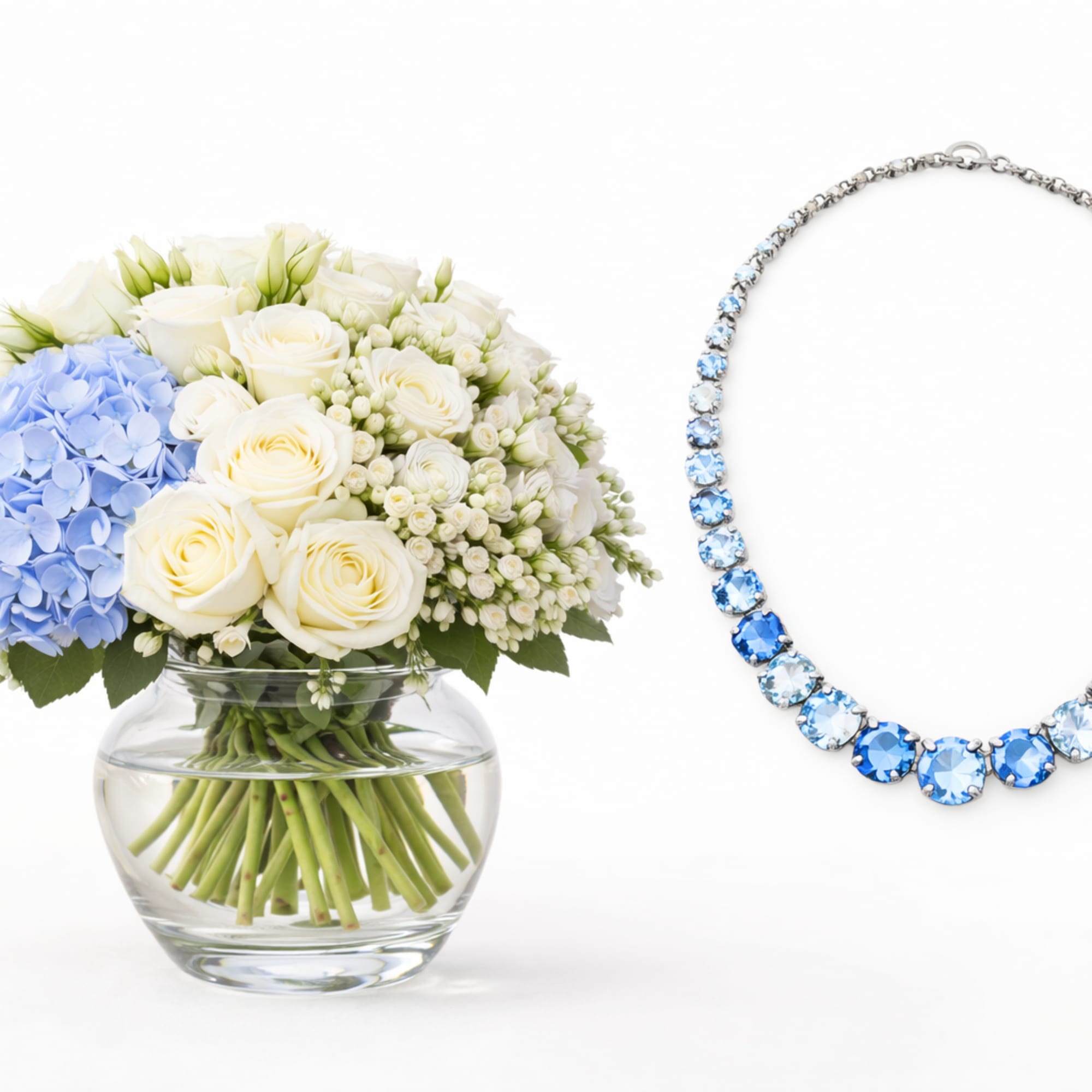 White roses and blue hydrangeas in a glass vase beside a blue gemstone necklace