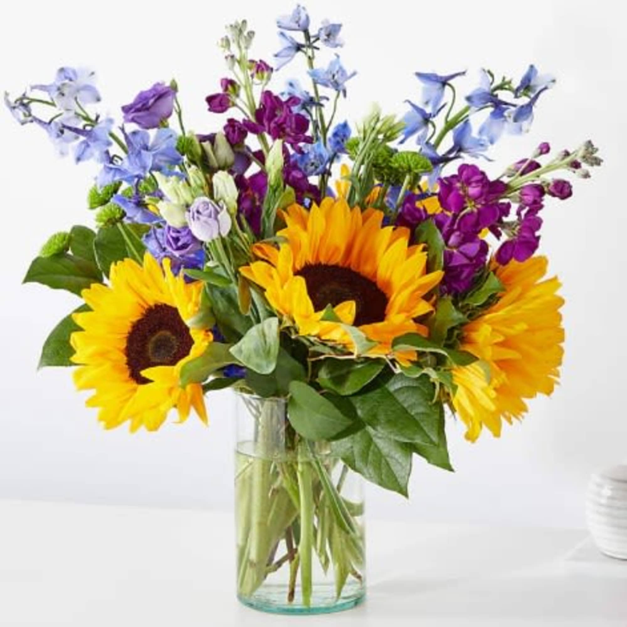 Clear vase of bright yellow sunflowers with purple and blue accent flowers