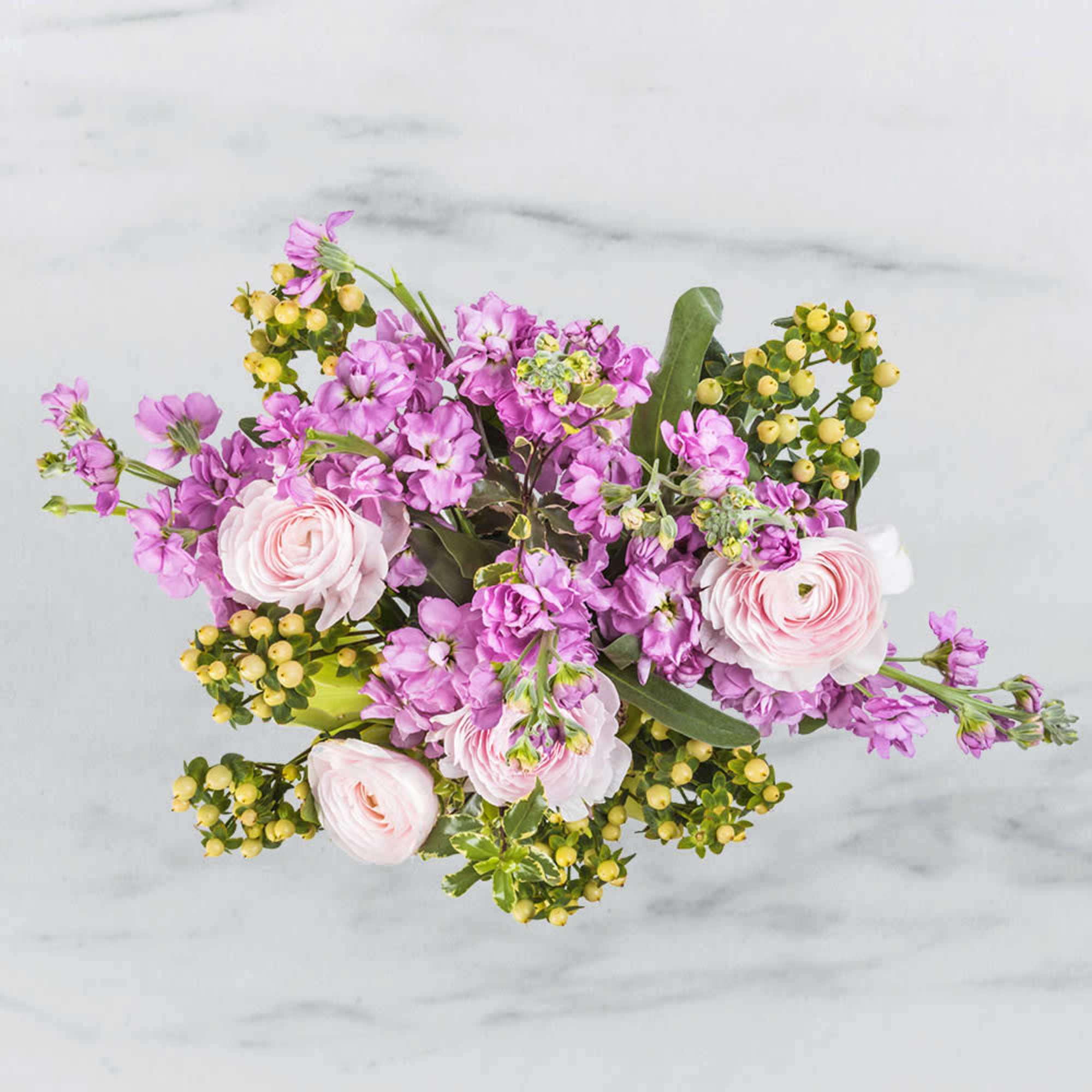 Top view of a bouquet of lavender stock and pale pink ranunculus with yellow berries on a light surface