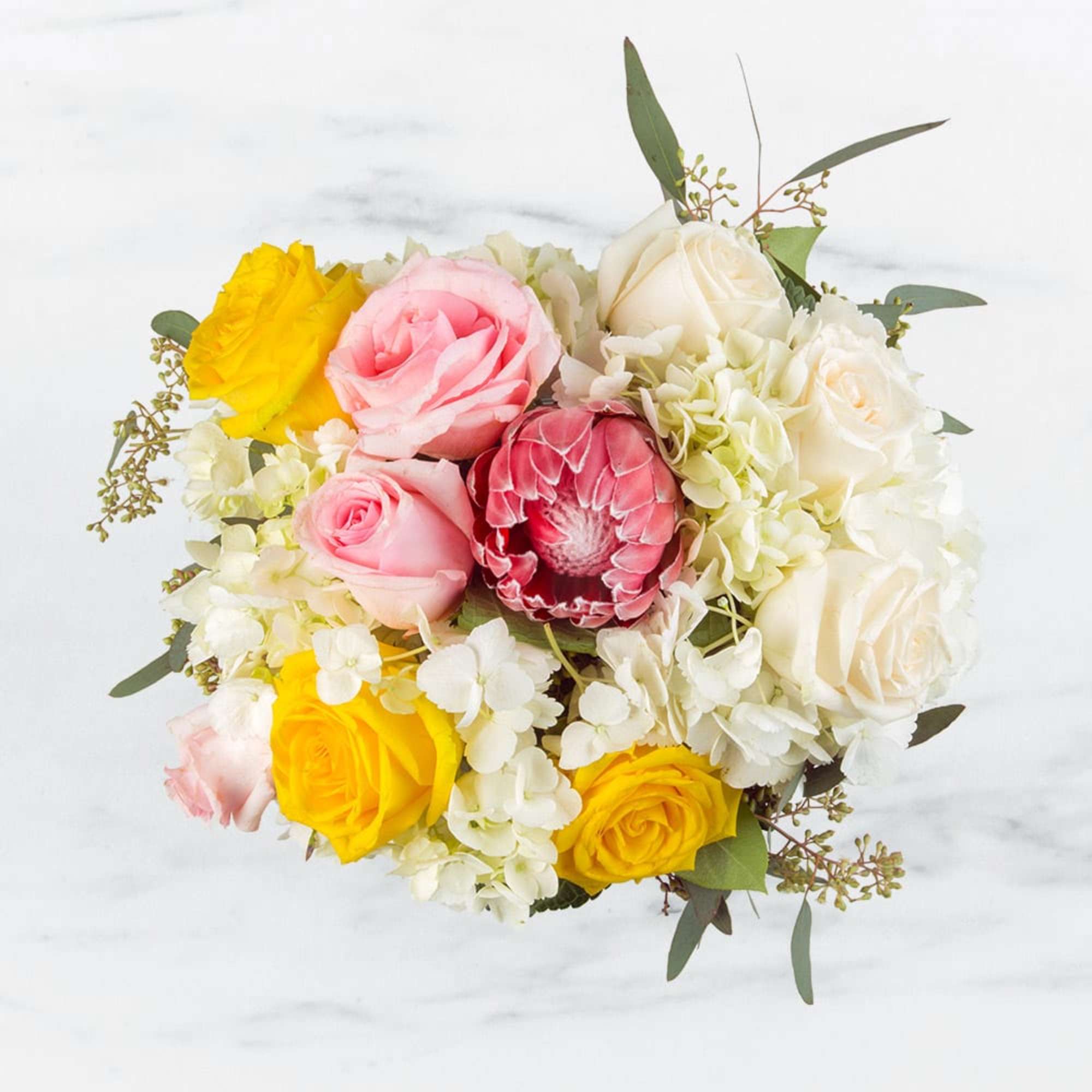 Round arrangement of pink and yellow roses, white hydrangeas, and a pink protea bloom