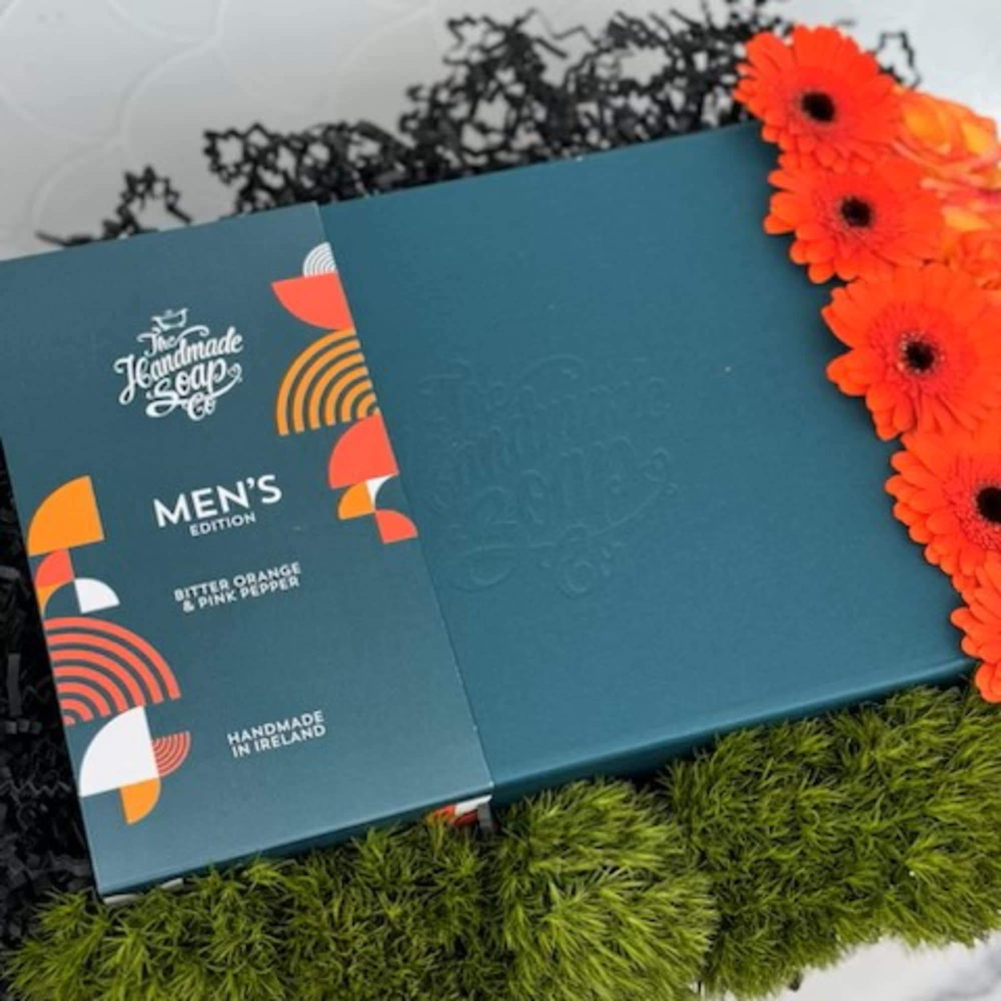 Teal men's soap gift box with bright orange gerbera daisies on moss and black shred