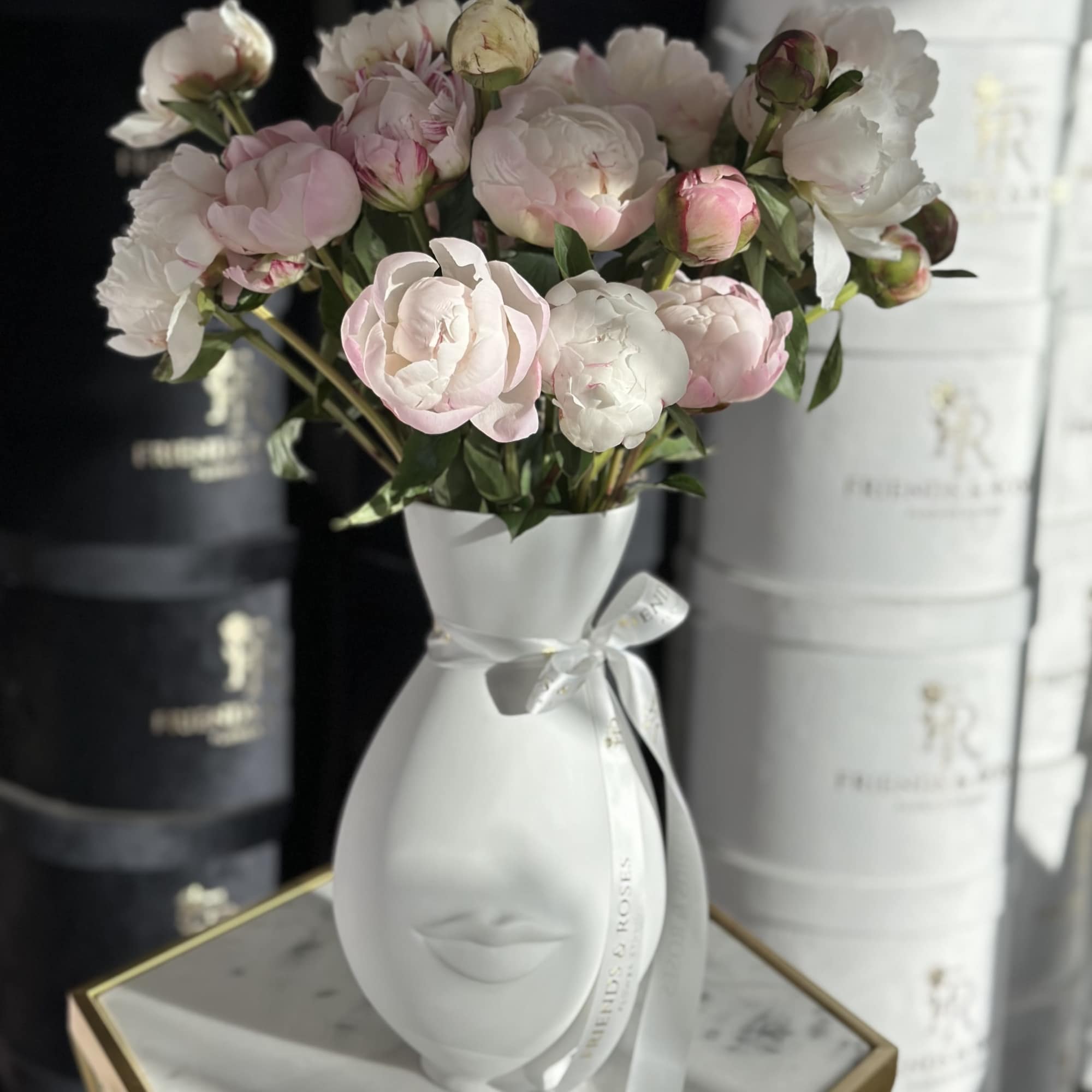 Pink and white peonies arranged in a white face-shaped vase with a ribbon