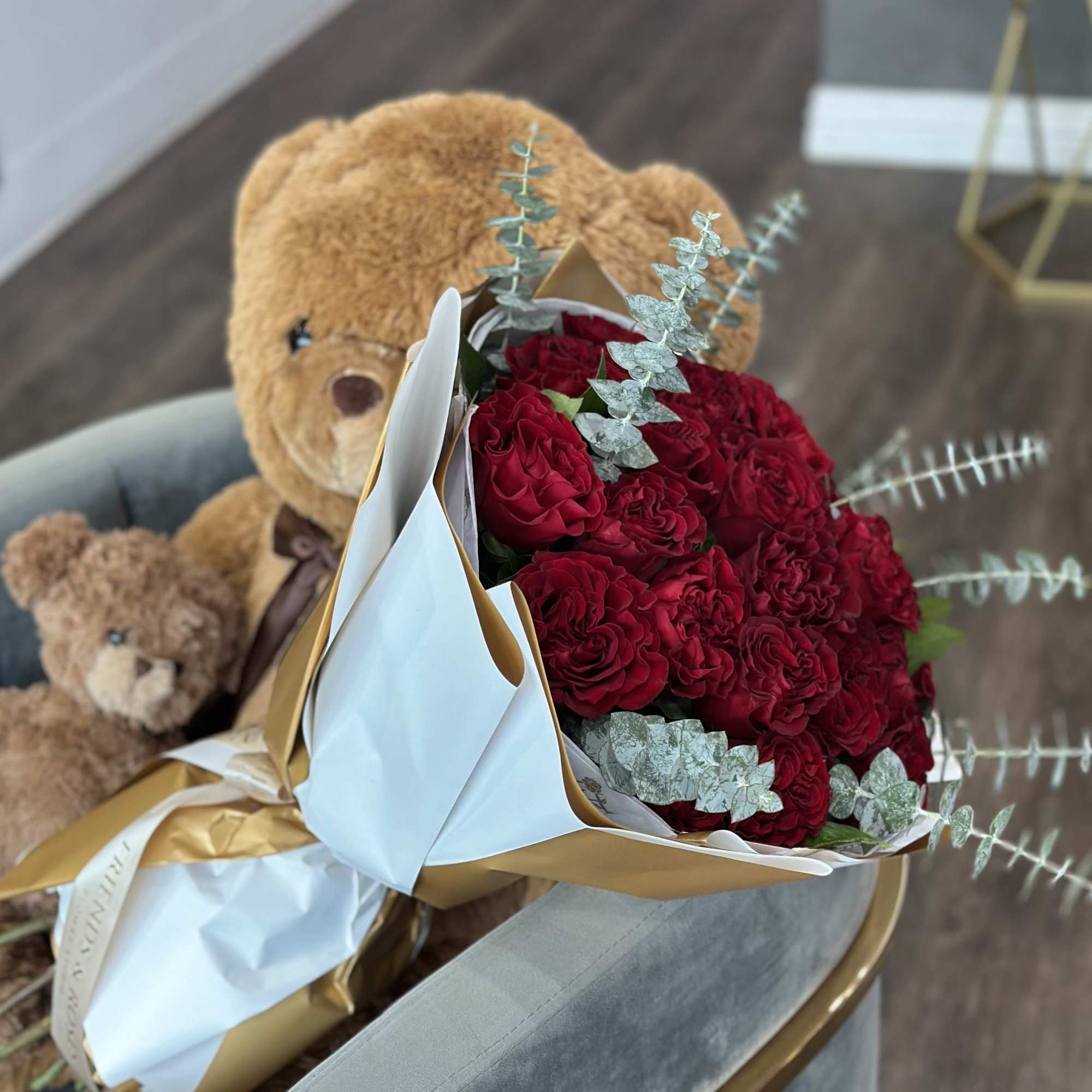 Bouquet of red roses wrapped in white and gold paper with a teddy bear behind it