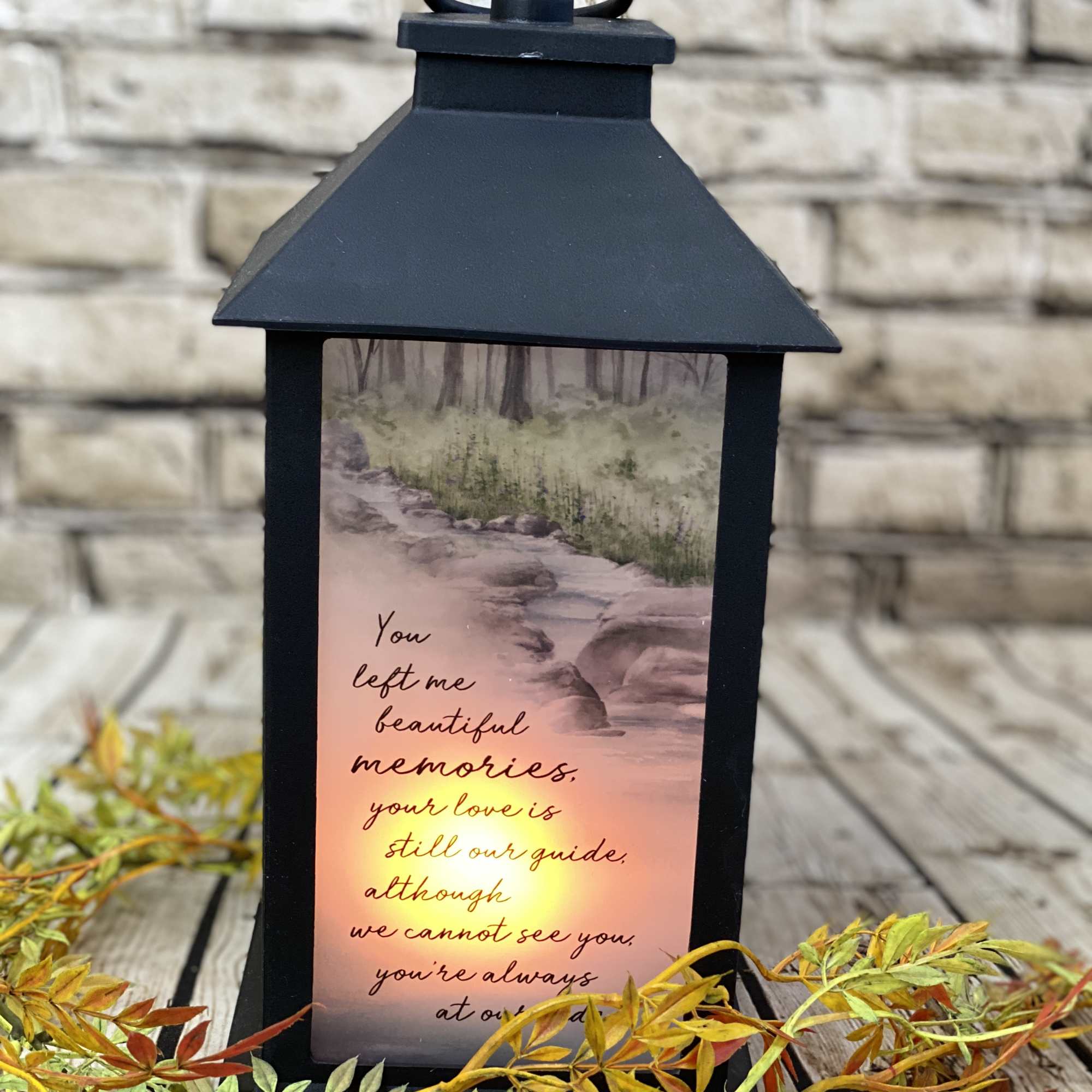 Black memorial lantern with glowing nature scene and remembrance verse, surrounded by autumn-colored decorative foliage.