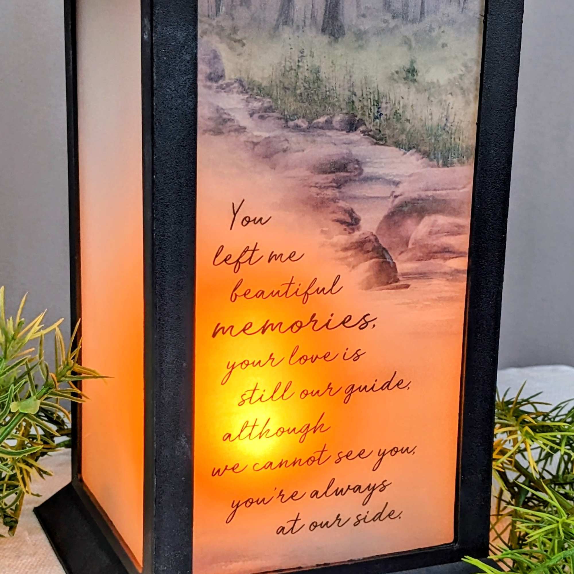 Black memorial lantern with warm glow and sentimental verse over a forest path scene