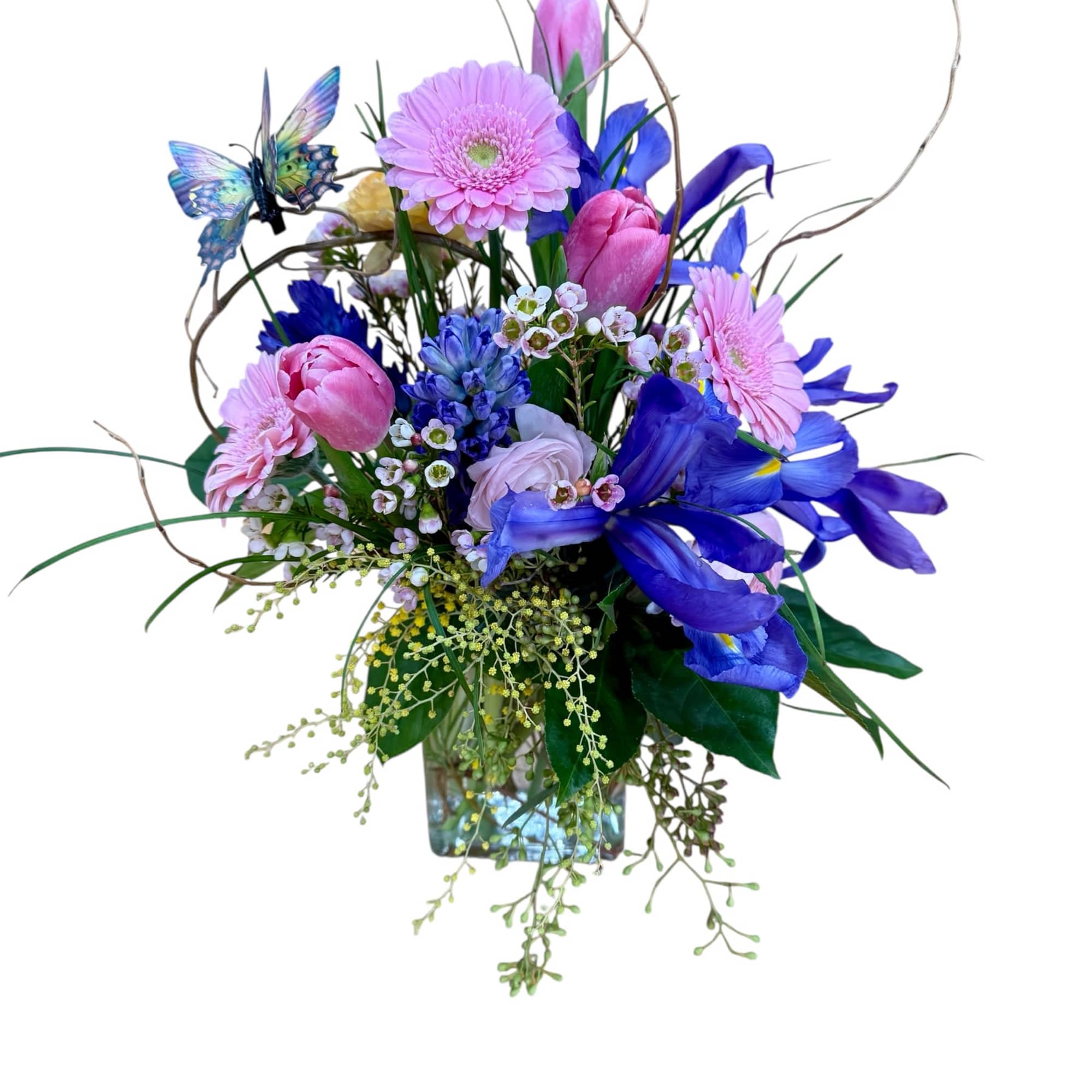 Tall arrangement of pink gerbera daisies, tulips, and blue irises in a glass cube with a butterfly accent